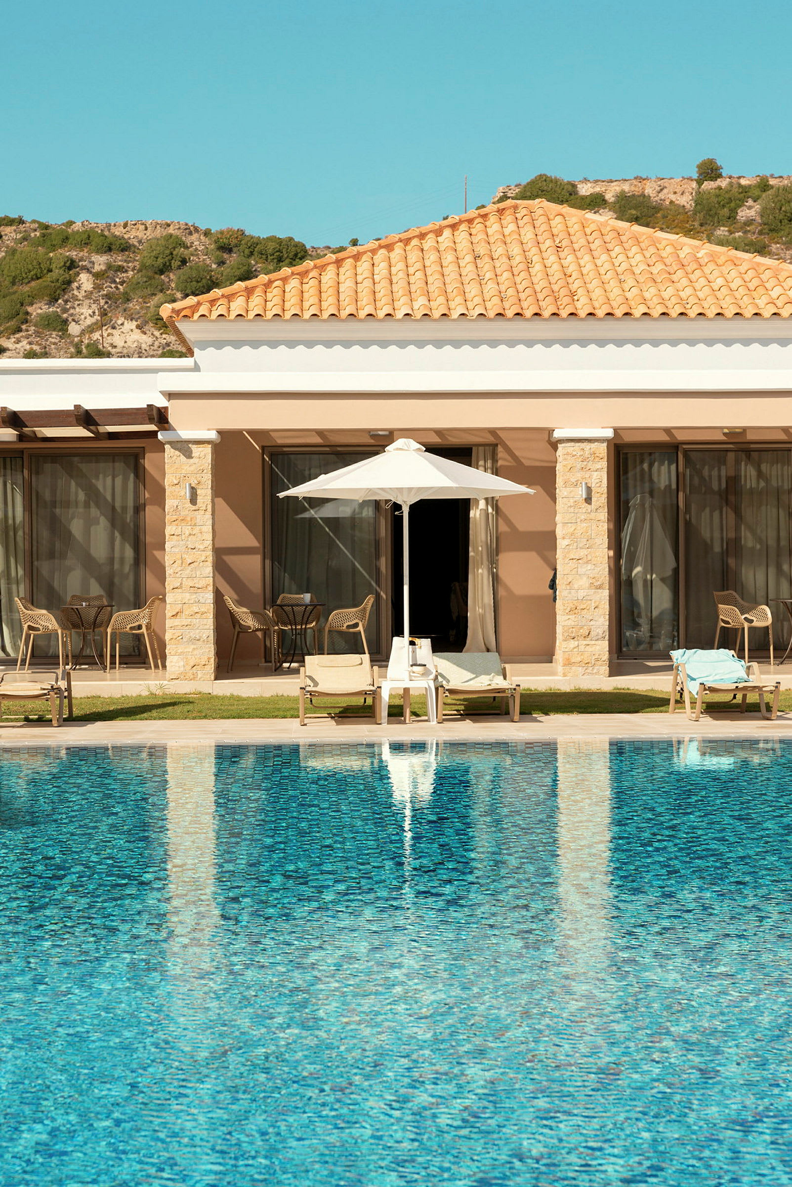 Pool foran hotelbygning med solsenge og parasol. (AI-genereret tekst)