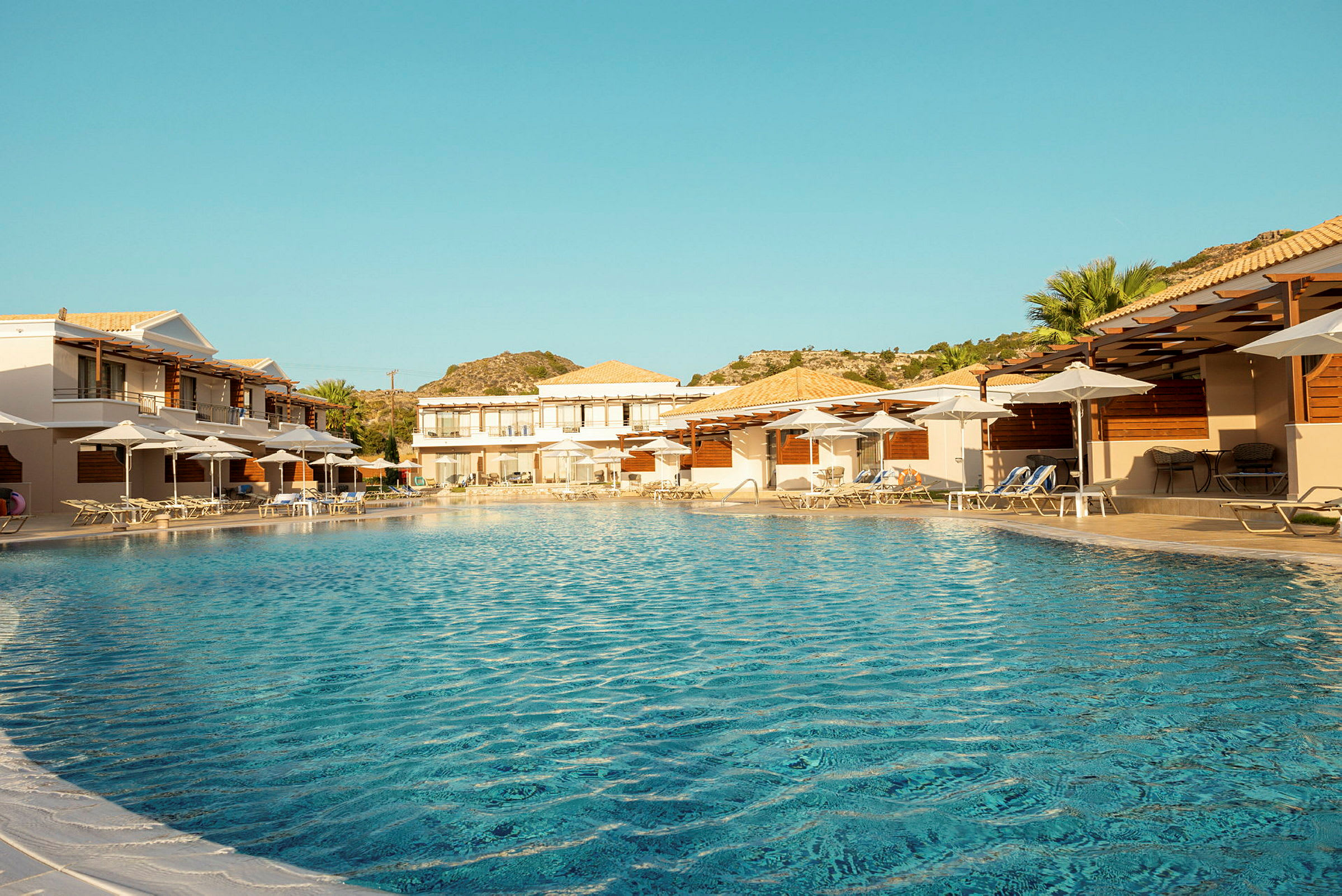 Billedet viser en swimmingpool omgivet af liggestole og parasoller ved hotelbygninger. (AI-genereret tekst)