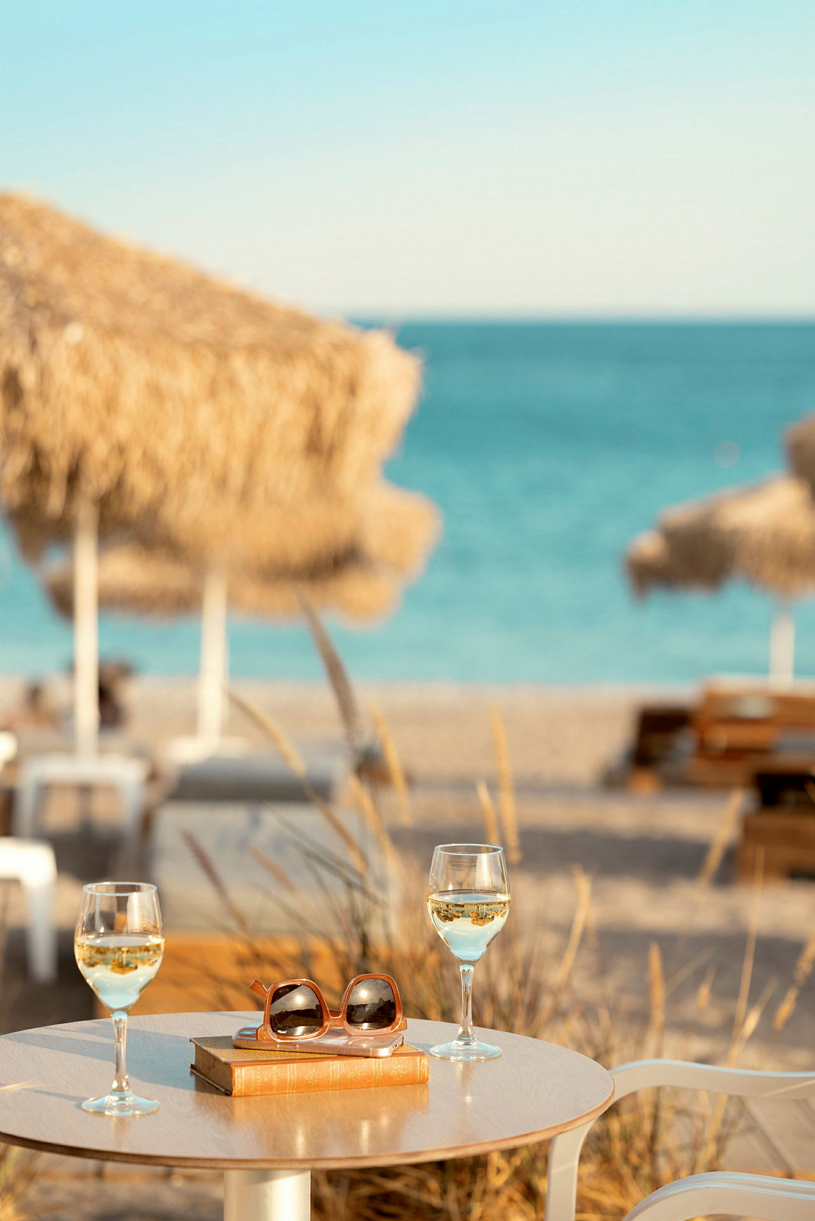 Bord med to glas hvidvin, en bog og solbriller ved en strand med parasoller og hav i baggrunden. (AI-genereret tekst)