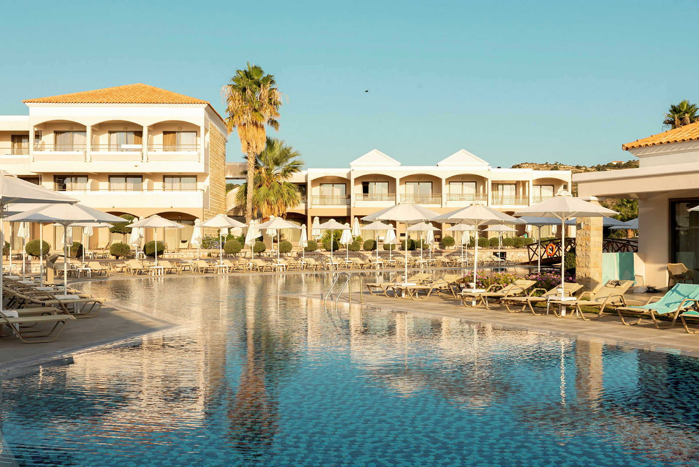 Hotelpool med liggestole og parasoller, omgivet af bygninger og palmer. (AI-genereret tekst)