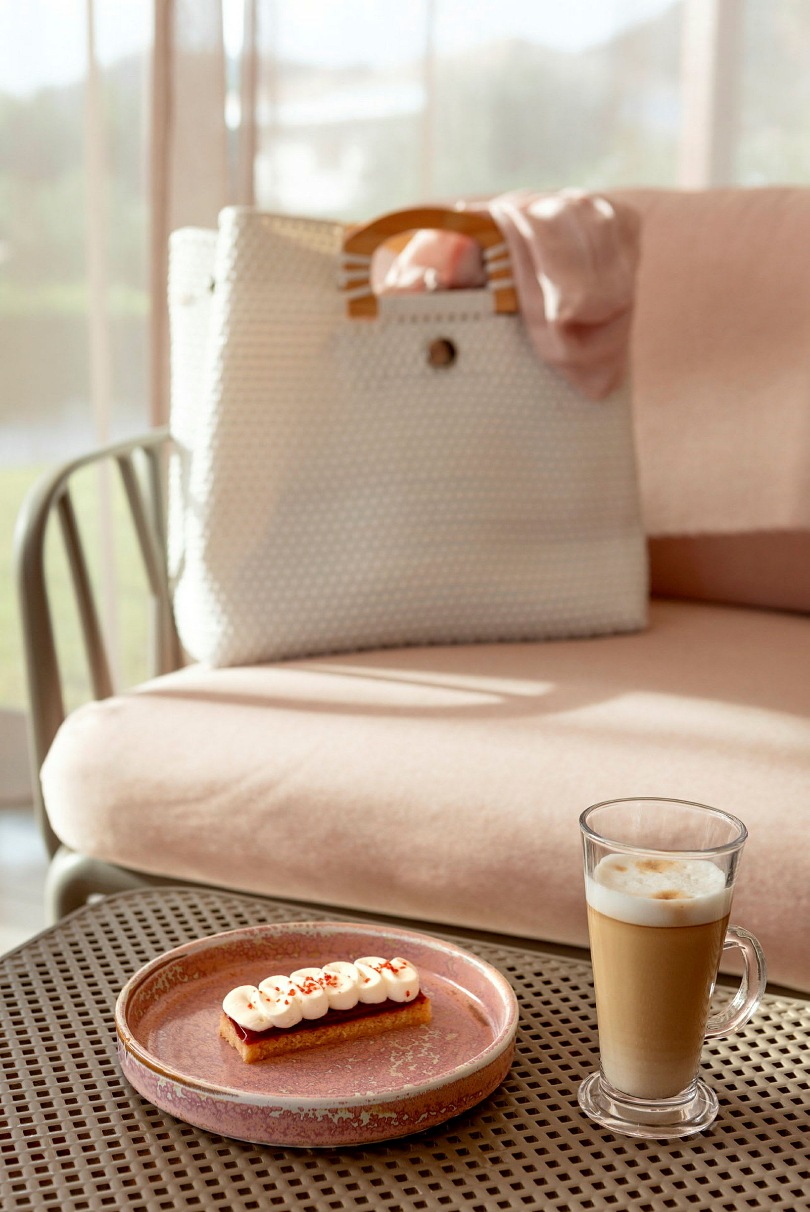 En beige taske med træhåndtag på en rosa siddeplads med et glas latte og en dessert på en tallerken i forgrunden. (AI-genereret tekst)