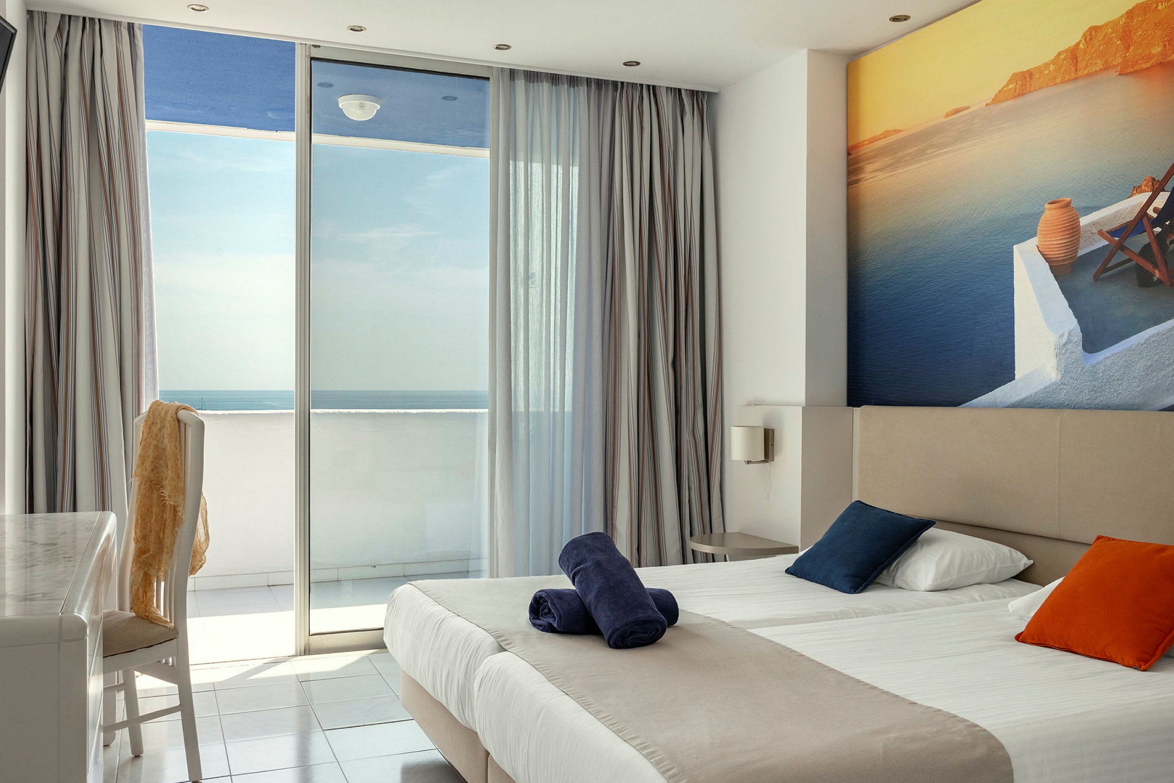 Hotelværelse med dobbeltseng, farverige puder og balkon med havudsigt. (AI-genereret tekst)