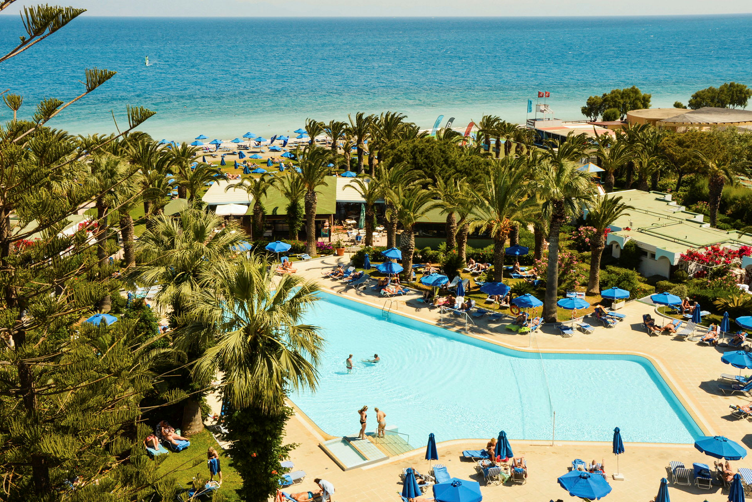 En pool omgivet af liggestole og palmer, med en sandstrand og havet i baggrunden. (AI-genereret tekst)