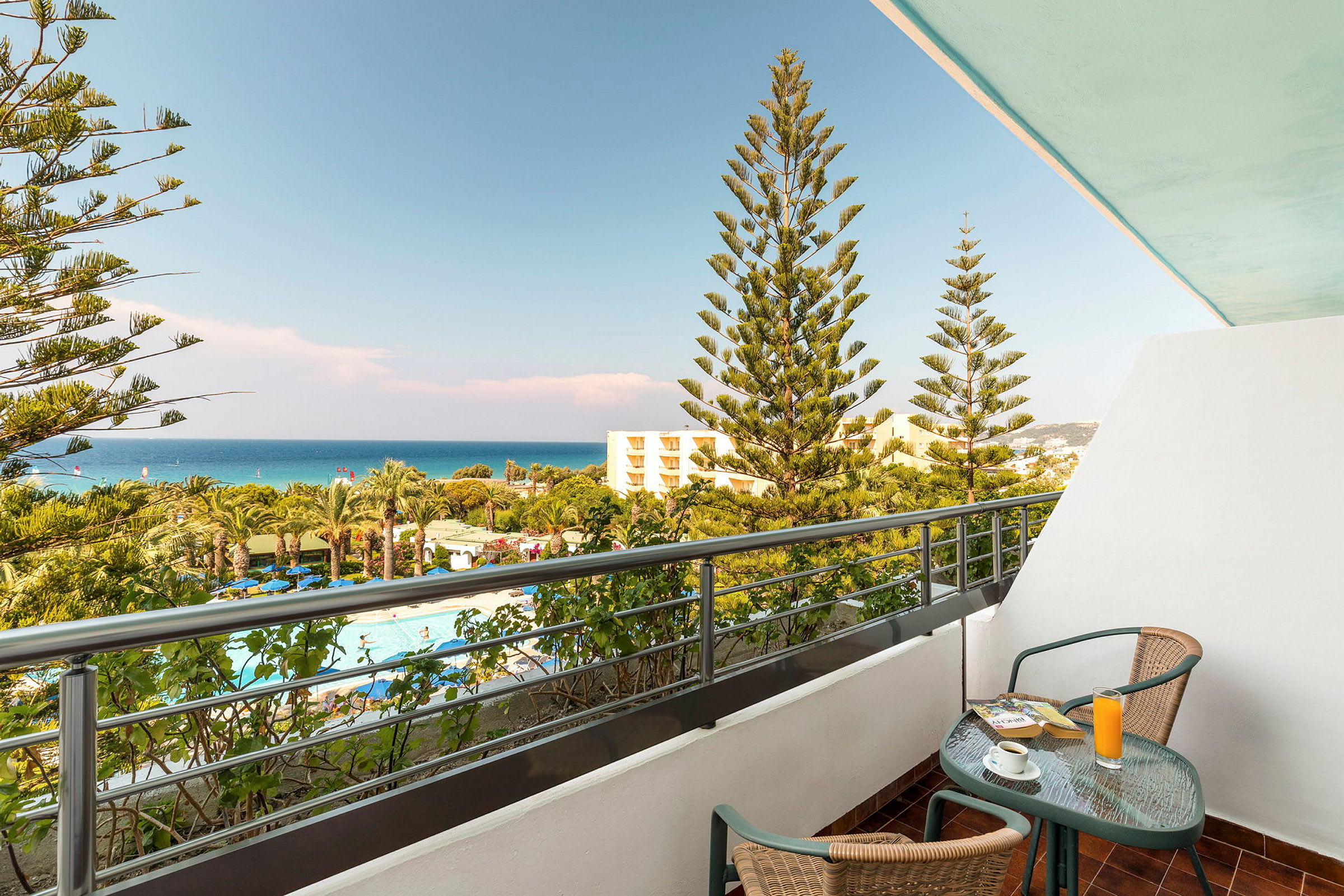 En balkon med et bord og to stole. Udsigt over en pool og frodigt område med palmer samt havet i baggrunden. (AI-genereret tekst)