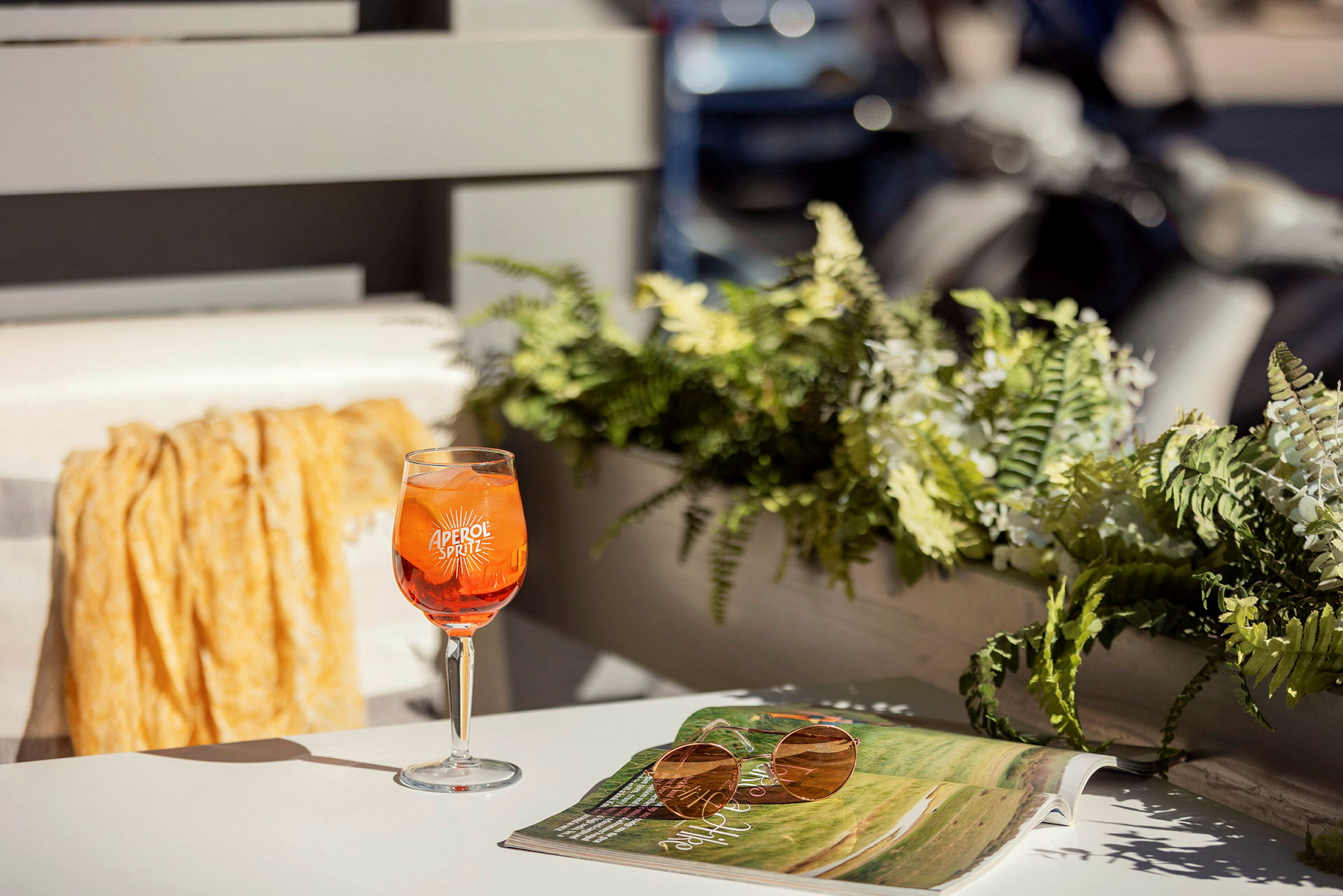 Et glas Aperol Spritz står på et bord ved siden af en solbrille og et åbent magasin. I baggrunden ses planter og dele af en altan. (AI-genereret tekst)