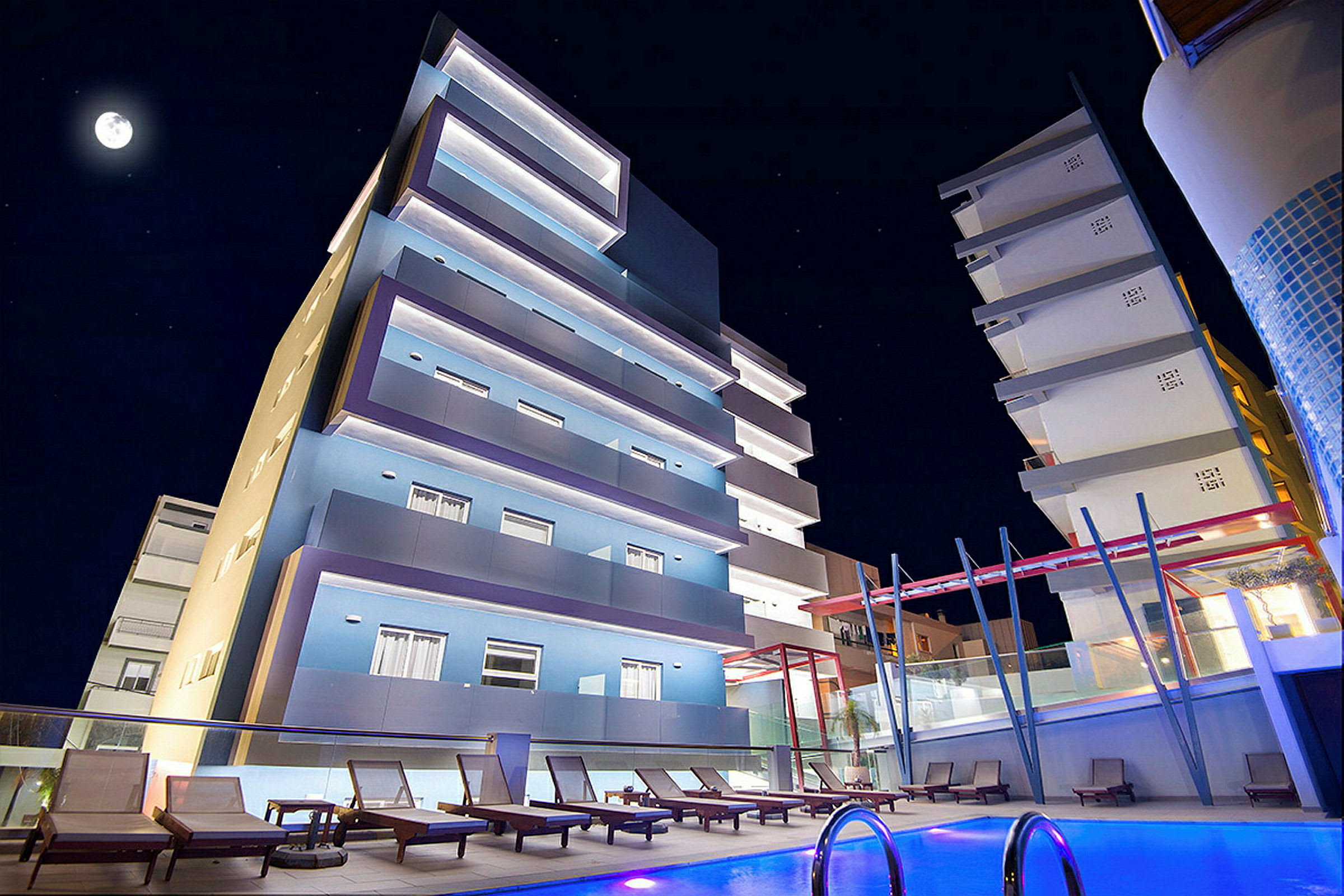 Et natbillede af et moderne hotel med oplyste facader, en pool i forgrunden og liggestole omkring. (AI-genereret tekst)