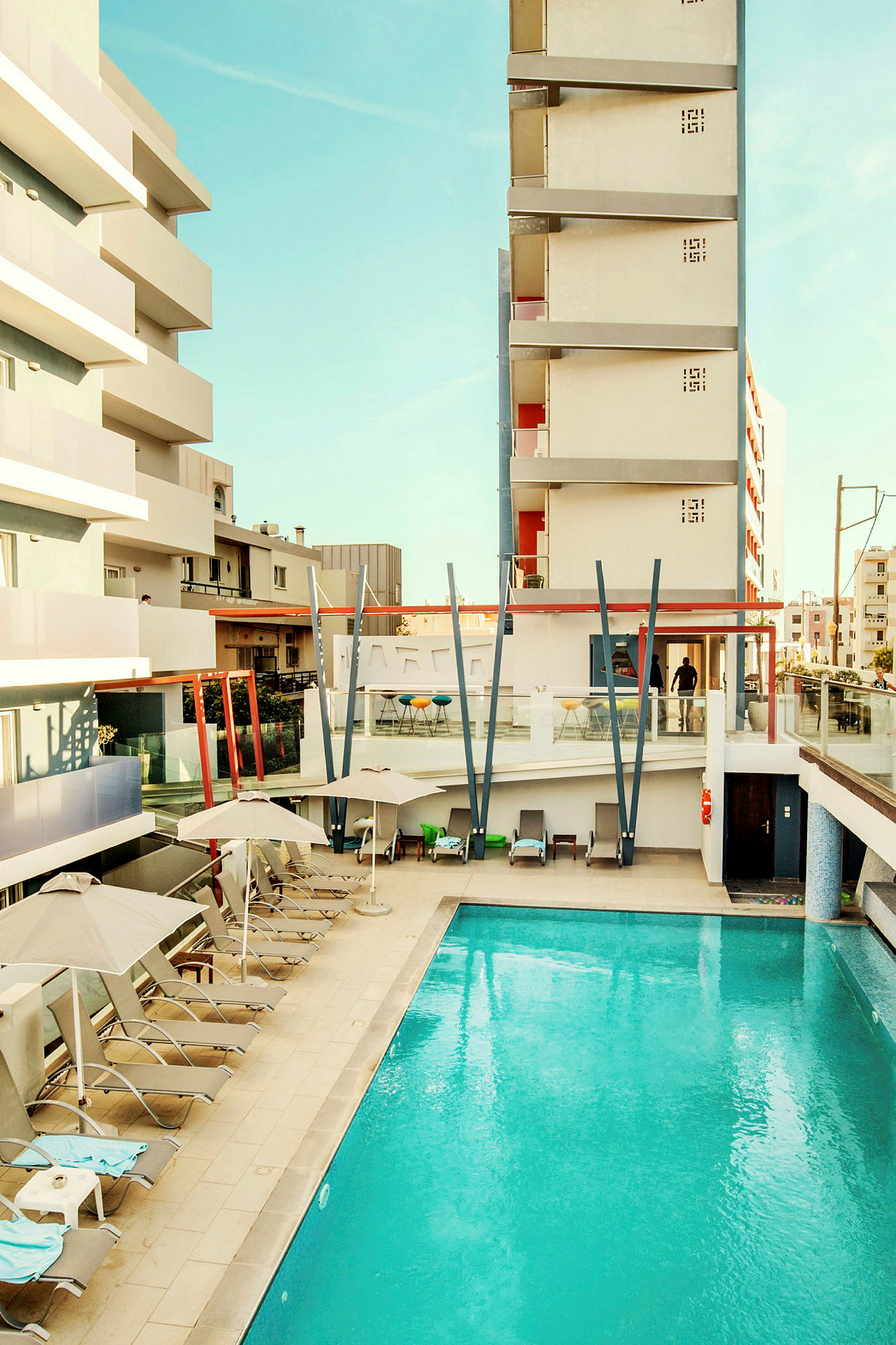 Billede af et hotelområde med pool og liggestole, omgivet af bygninger. (AI-genereret tekst)