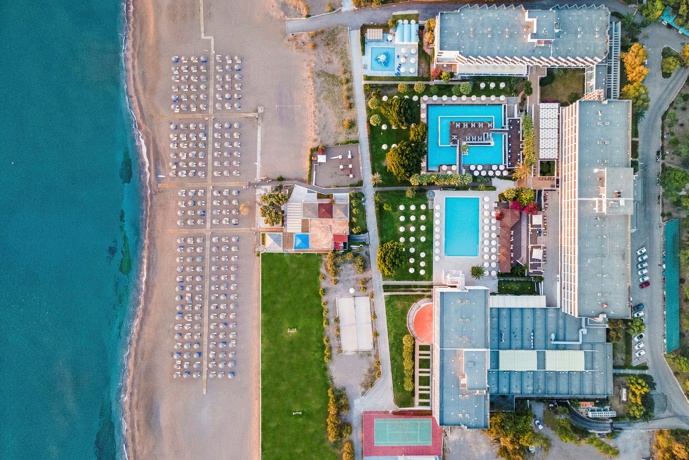 Luftfoto af en strand med liggestole og parasoller ved havet samt et hotelområde med pools og bygninger. (AI-genereret tekst)