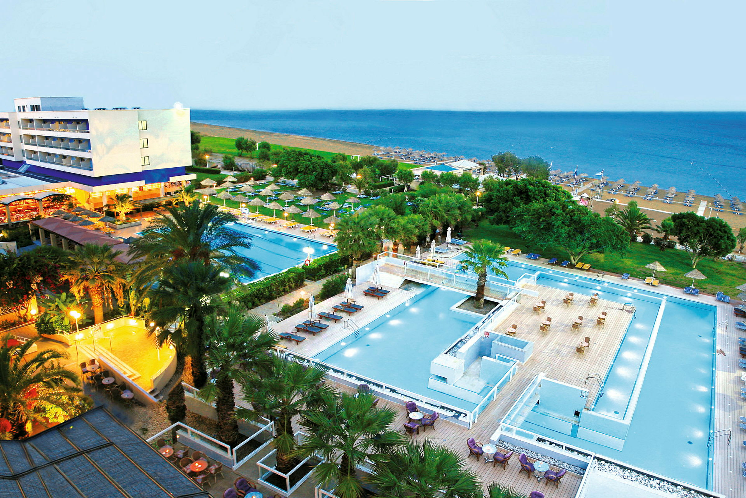 Udsigt over et hotelområde med swimmingpools og grønne områder, med en sandstrand og havet i baggrunden. (AI-genereret tekst)
