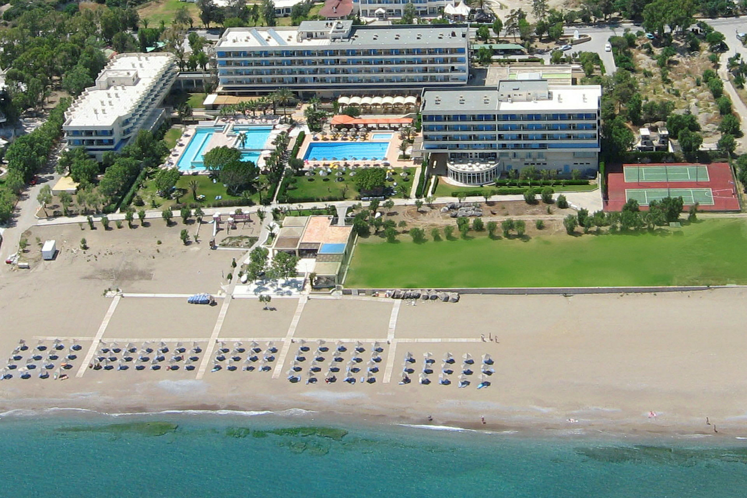 Luftperspektiv af et hotelkompleks med swimmingpools, grønne områder og tennisbaner nær en sandstrand. (AI-genereret tekst)
