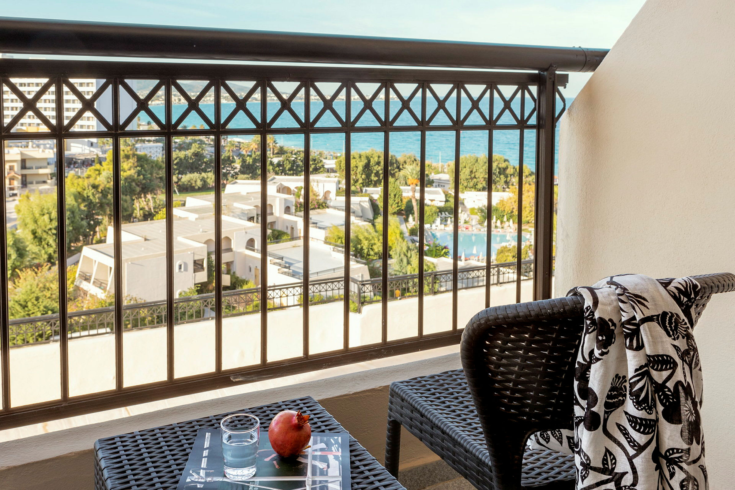 En balkon med udsigt over hotelområdet og havet. På altanbordet står et glas vand og en granatæble. På en stol ligger et mønstret sjal. (AI-genereret tekst)