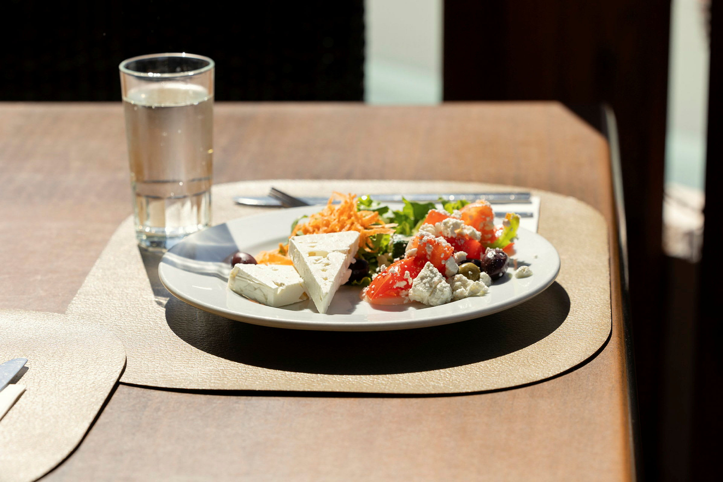 En tallerken med salat, grøntsager og fetaost samt et glas vand på et bord. (AI-genereret tekst)