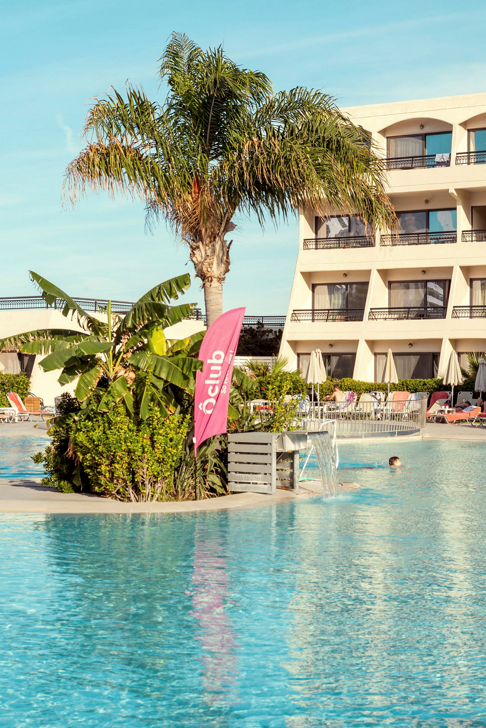 Udendørs pool foran hotelbygning med palmer og bevoksning. (AI-genereret tekst)
