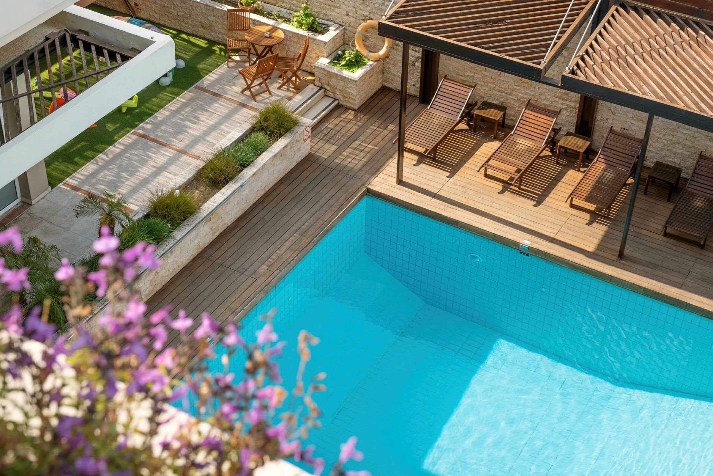 Udendørs swimmingpool med omkringliggende have og liggestole. Blomster i forgrunden. (AI-genereret tekst)