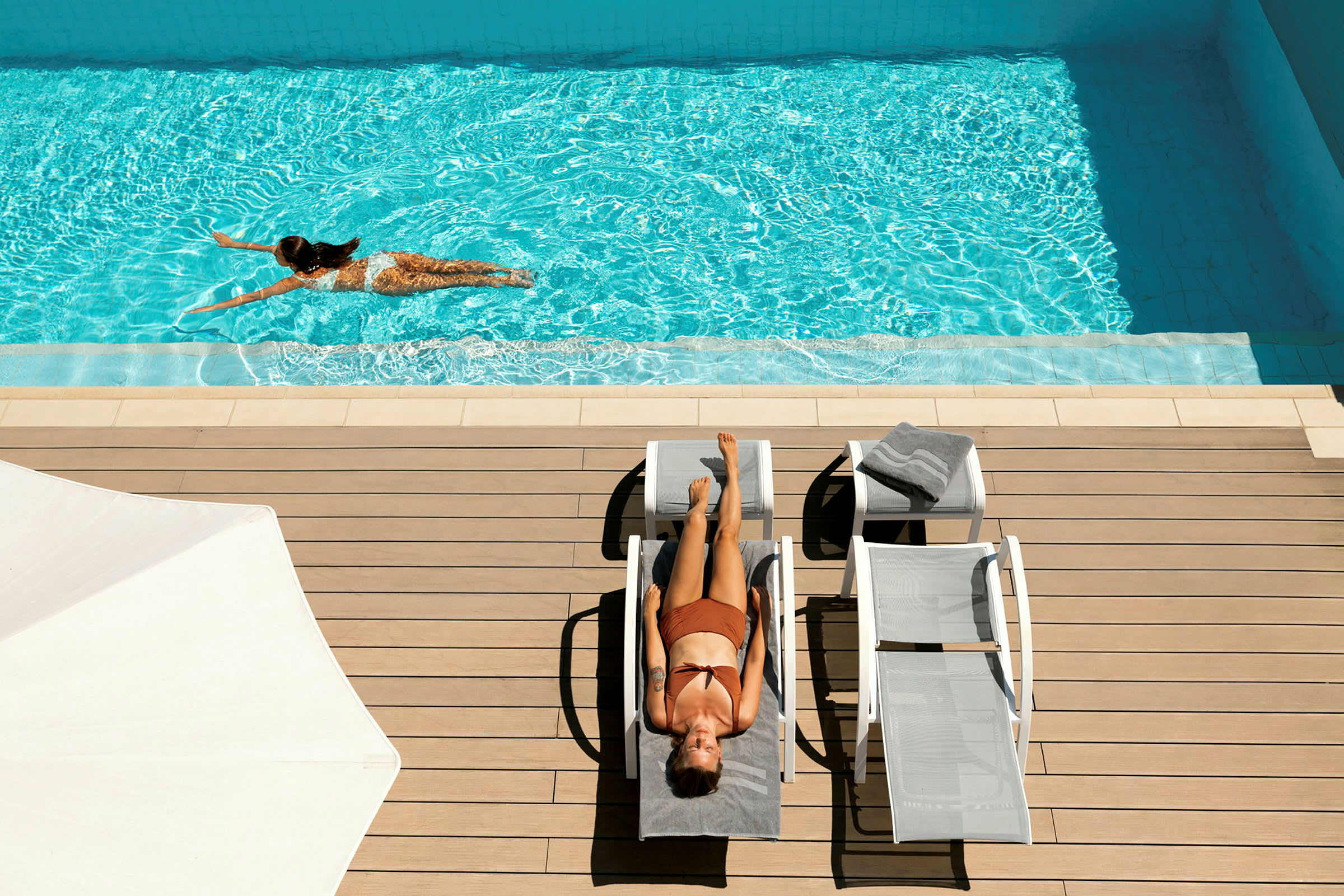 En kvinde ligger og solbader på en solvogn ved en pool. En anden kvinde svømmer i poolen. (AI-genereret tekst)