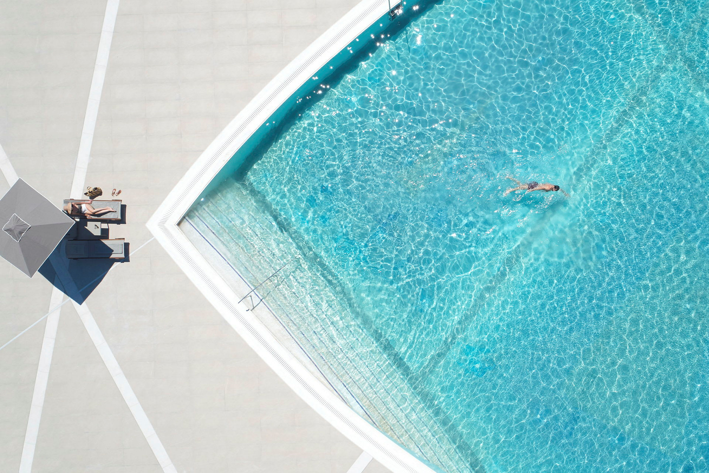 En pool med klart blåt vand, en person svømmer og en anden slapper af under en parasol ved poolkanten. (AI-genereret tekst)