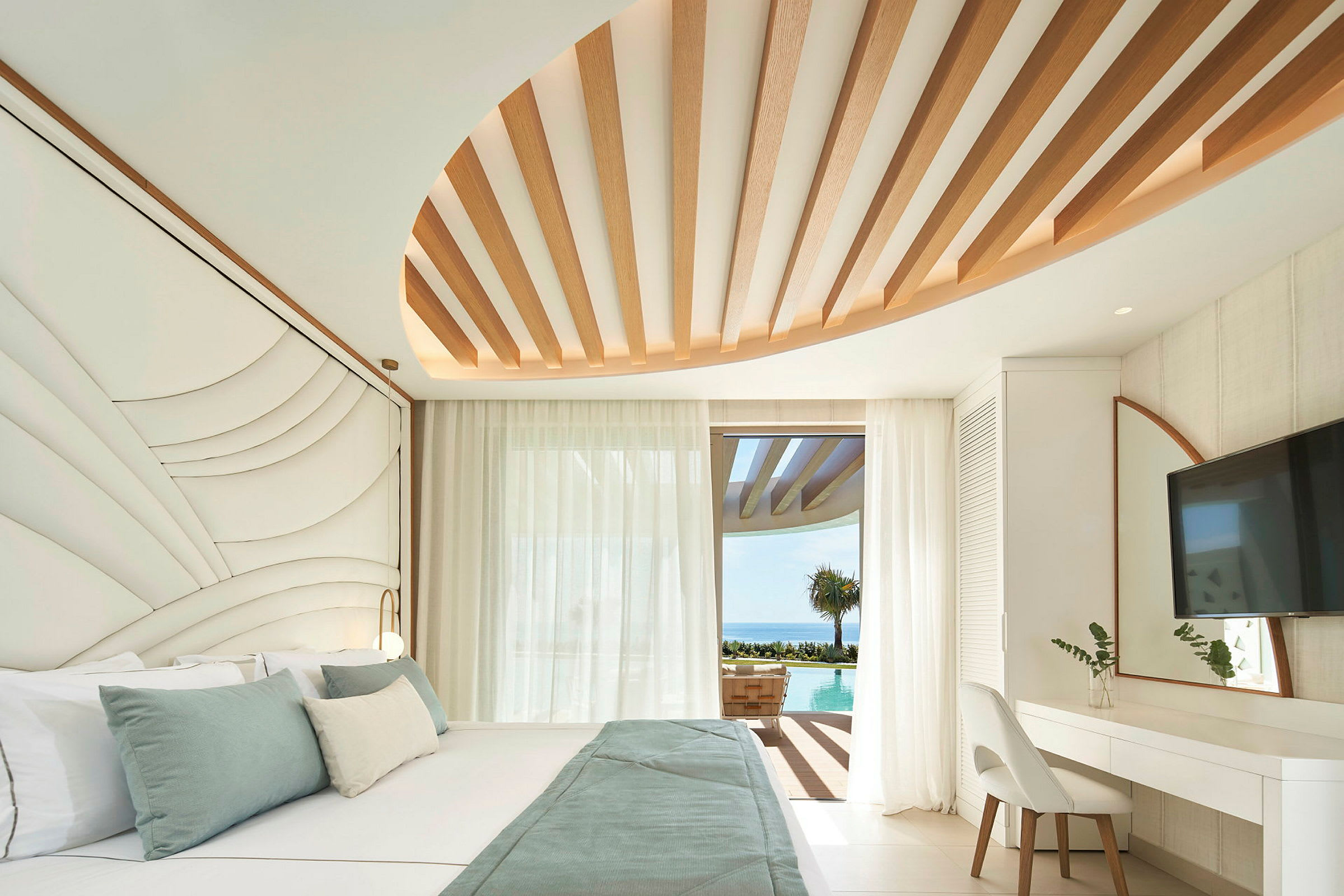 Et moderne hotelværelse med en stor seng, designede loftdetaljer og udsigt over en pool og havet gennem en glasdør. (AI-genereret tekst)