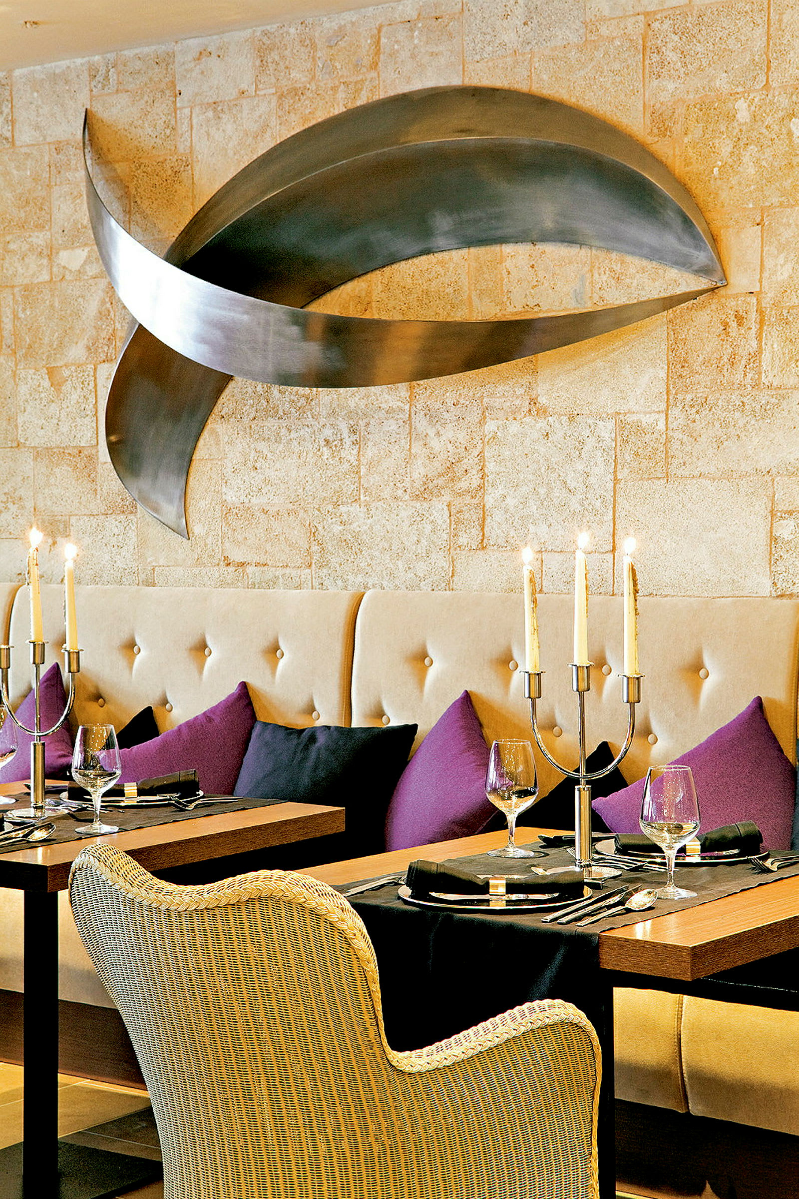 Et elegant restaurantbord med tallerkener, glas og lysestager, omgivet af polstrede sofaer og farvede puder, og en moderne kunstinstallation på væggen. (AI-genereret tekst)