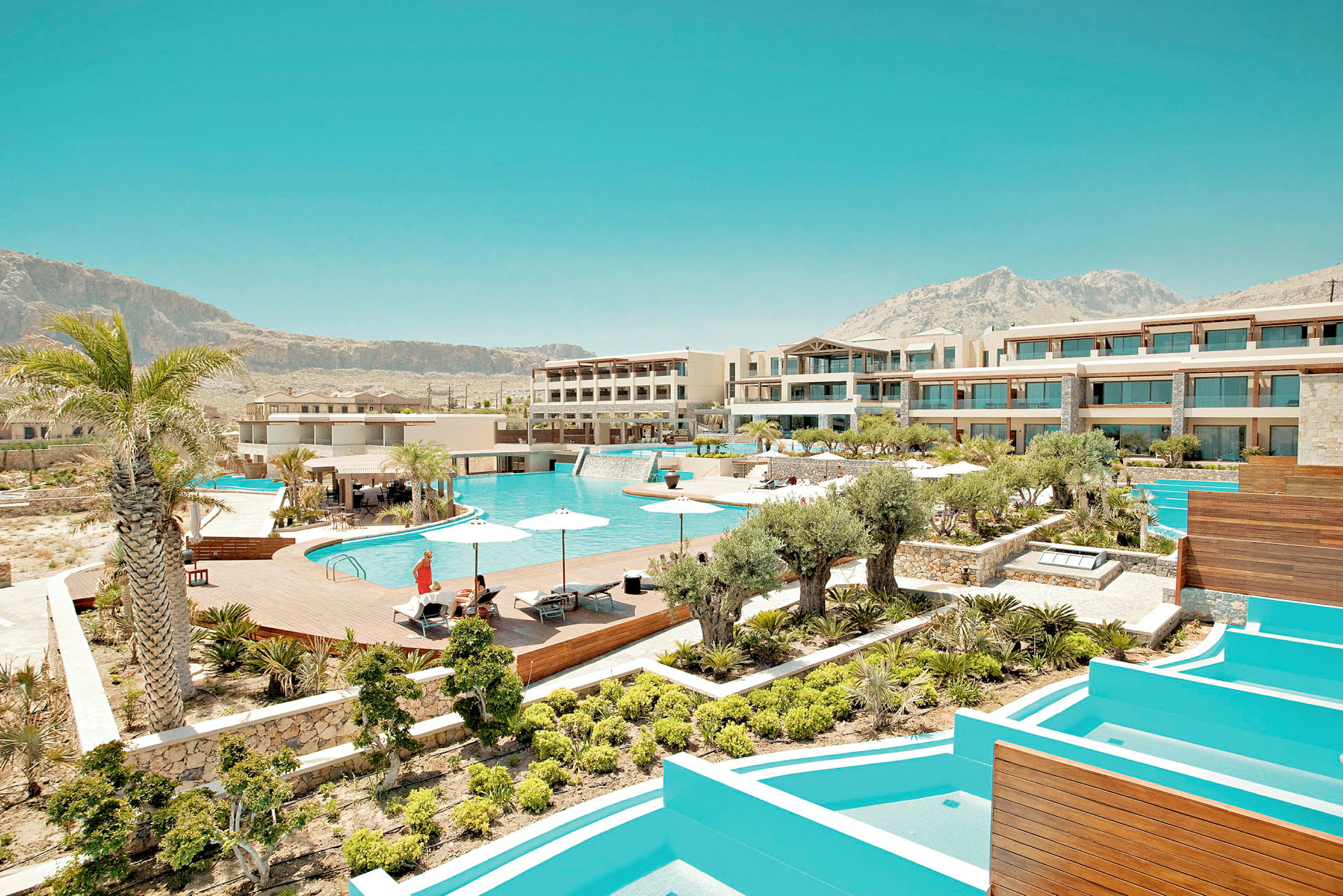 Et moderne resort med flere bygninger, en stor pool, parasoller og omgivelser med grønne områder. (AI-genereret tekst)