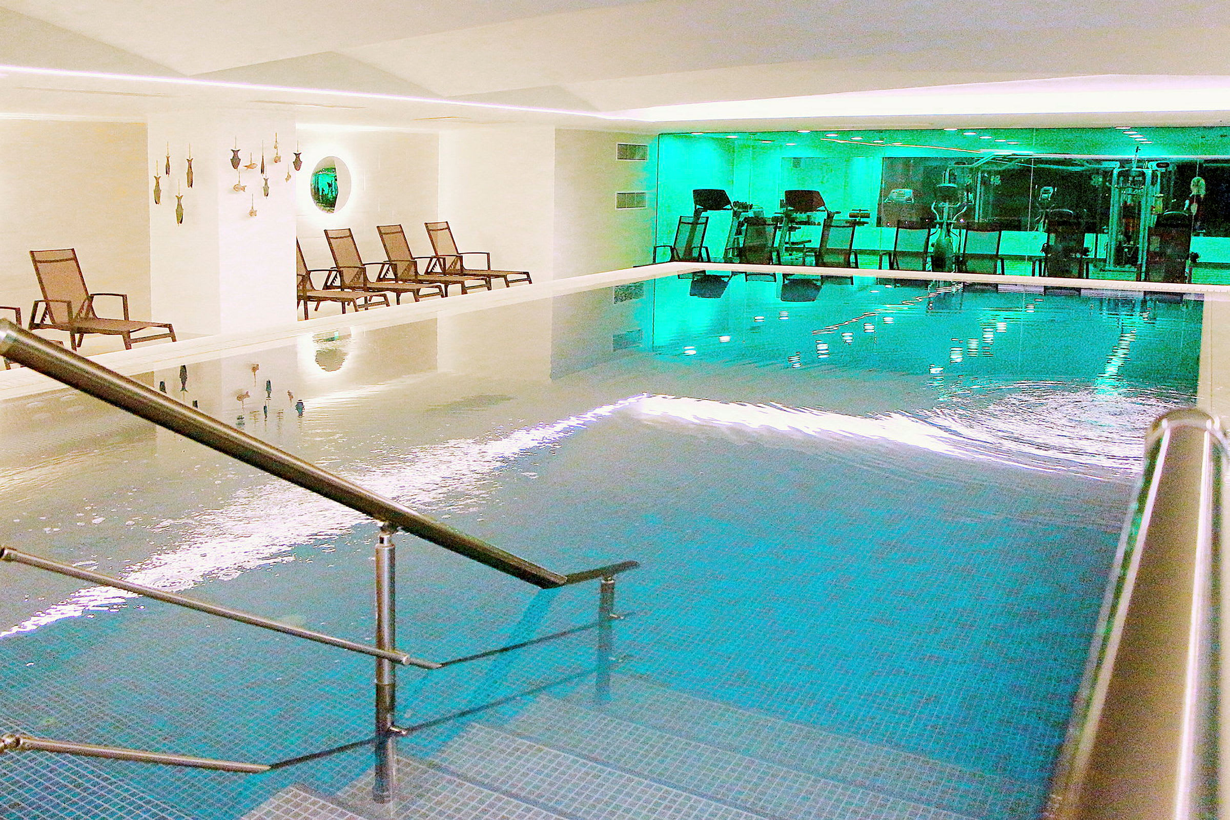 En indendørs pool med trapper og rækværk. Siddepladser og fitnessudstyr ses i baggrunden. (AI-genereret tekst)