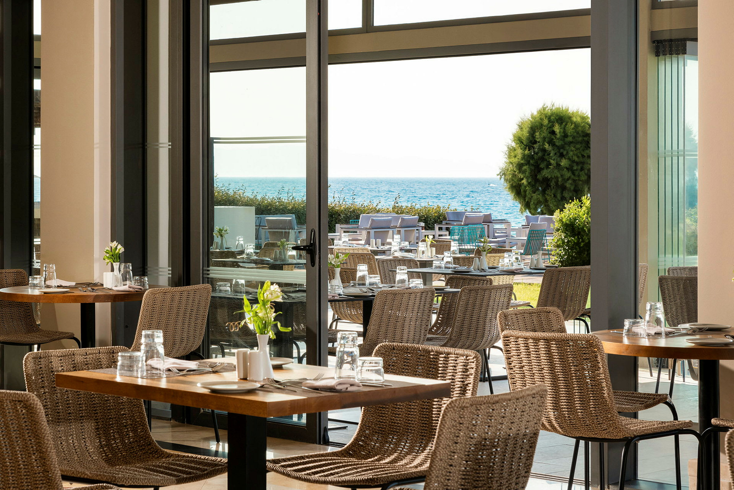 Udsigt over en moderne restaurant med flettestole og borde, med en have og havet i baggrunden. (AI-genereret tekst)