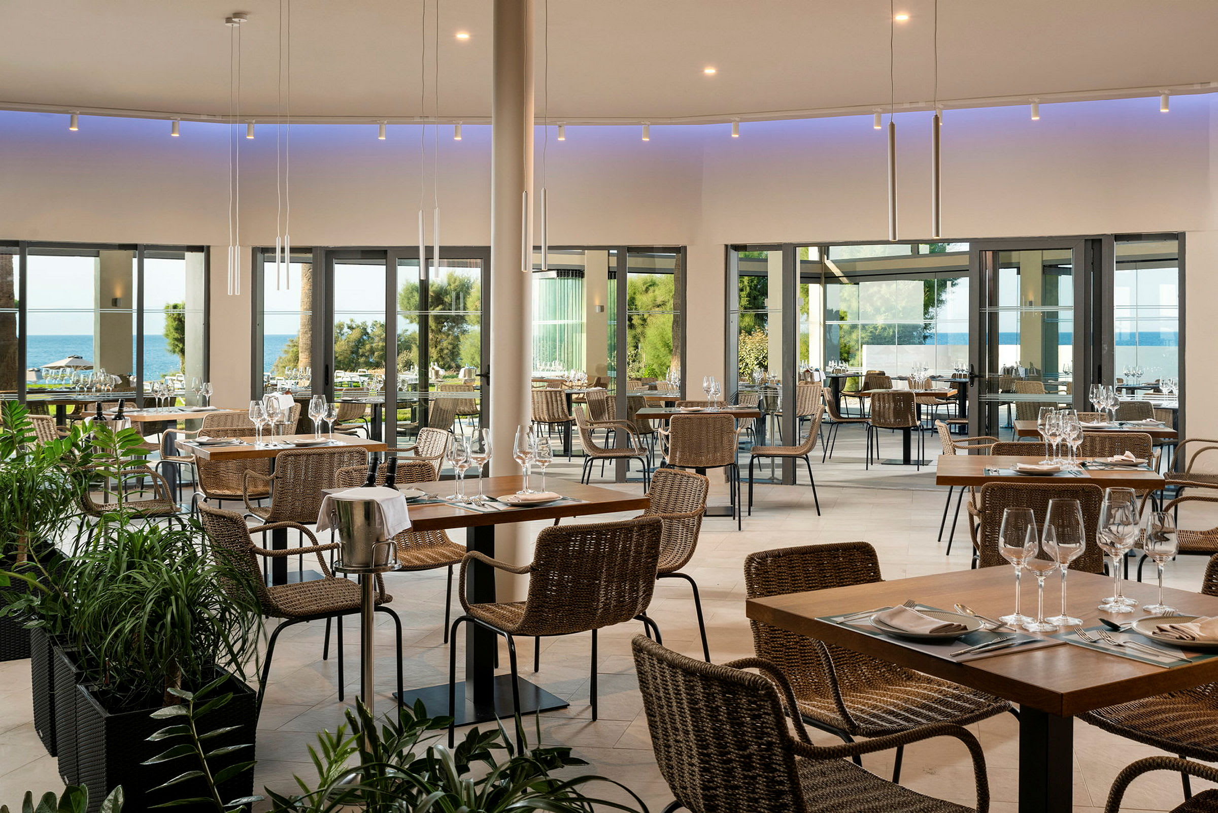 En restaurant med borde og stole, store glasvinduer med udsigt over havet og grøntområde. (AI-genereret tekst)