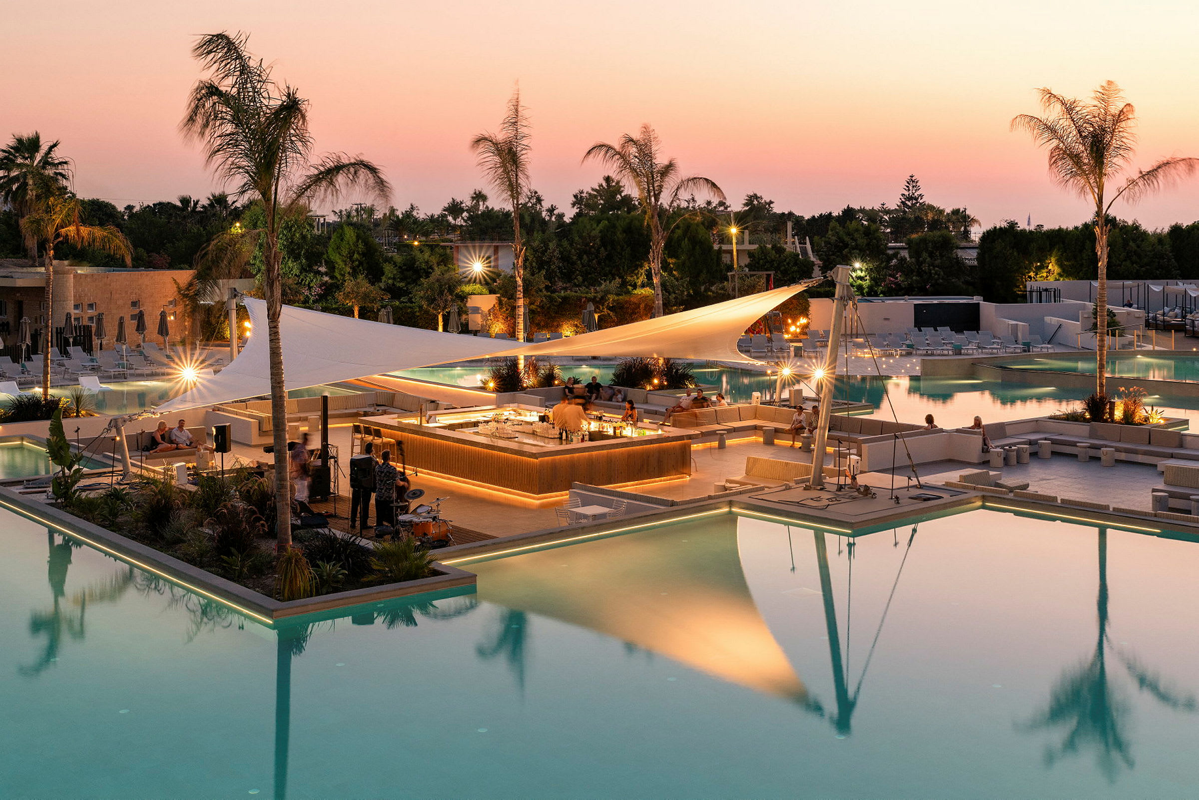Et ferieanlæg med oplyste swimmingpools og en central bar, placeret under et sejl ved solnedgang. (AI-genereret tekst)