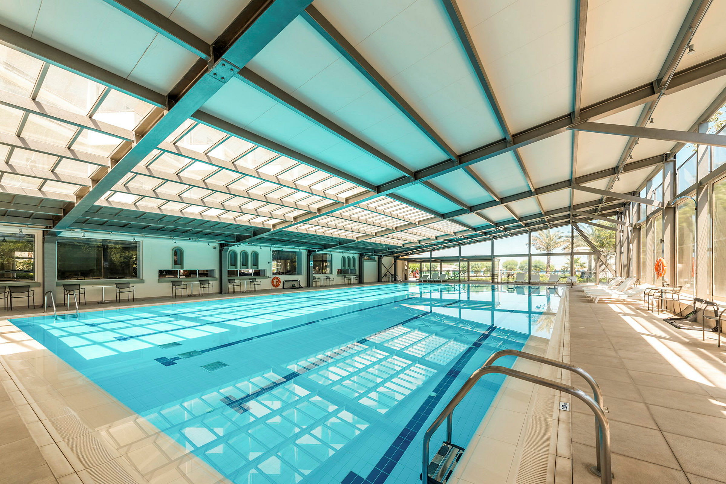 Indendørs poolområde med en stor pool, stole langs kanten og glasvægge, der slipper lys ind. (AI-genereret tekst)