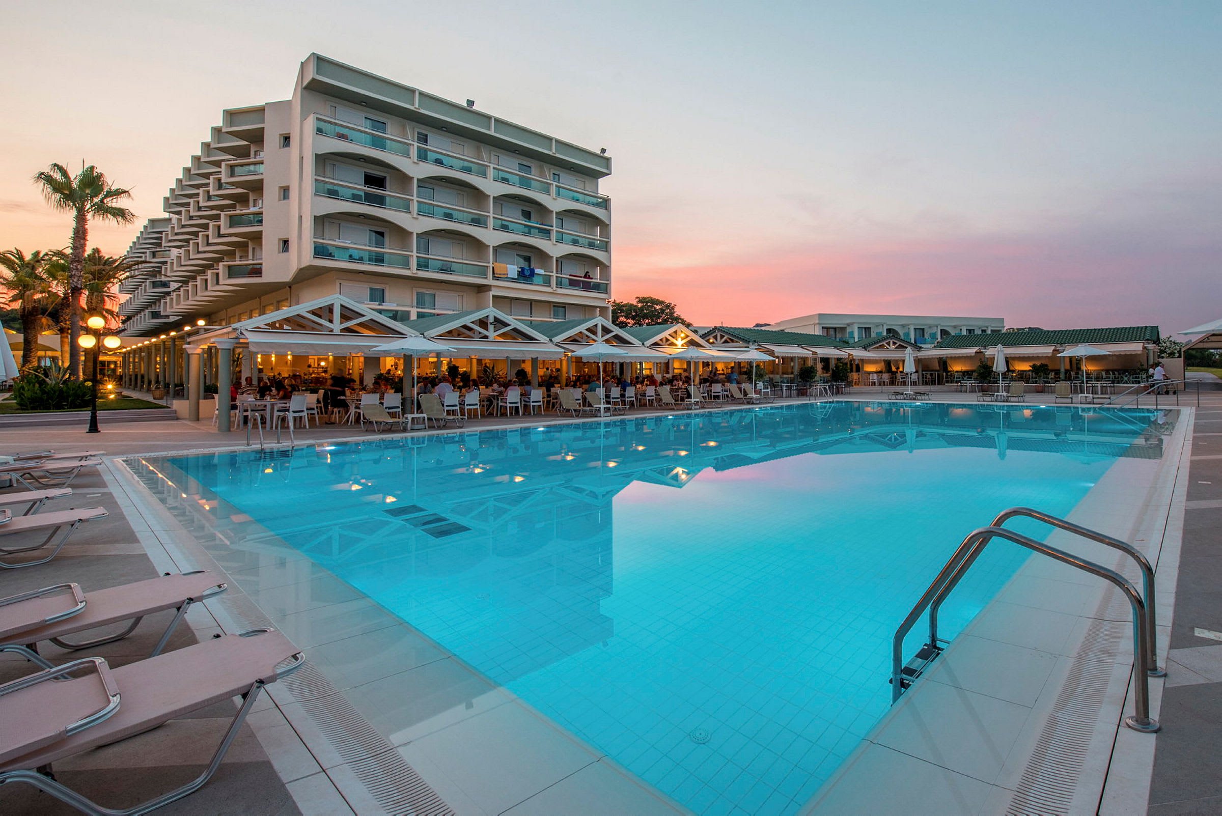 Et hotelbygning med altaner ved en stor swimmingpool omgivet af liggestole og en terrasse med siddepladser. (AI-genereret tekst)