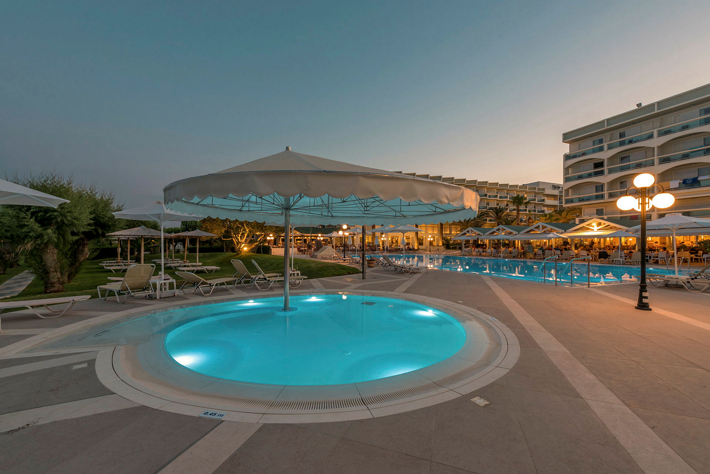 Hotellets poolområde om aftenen med parasoller og solstole. (AI-genereret tekst)