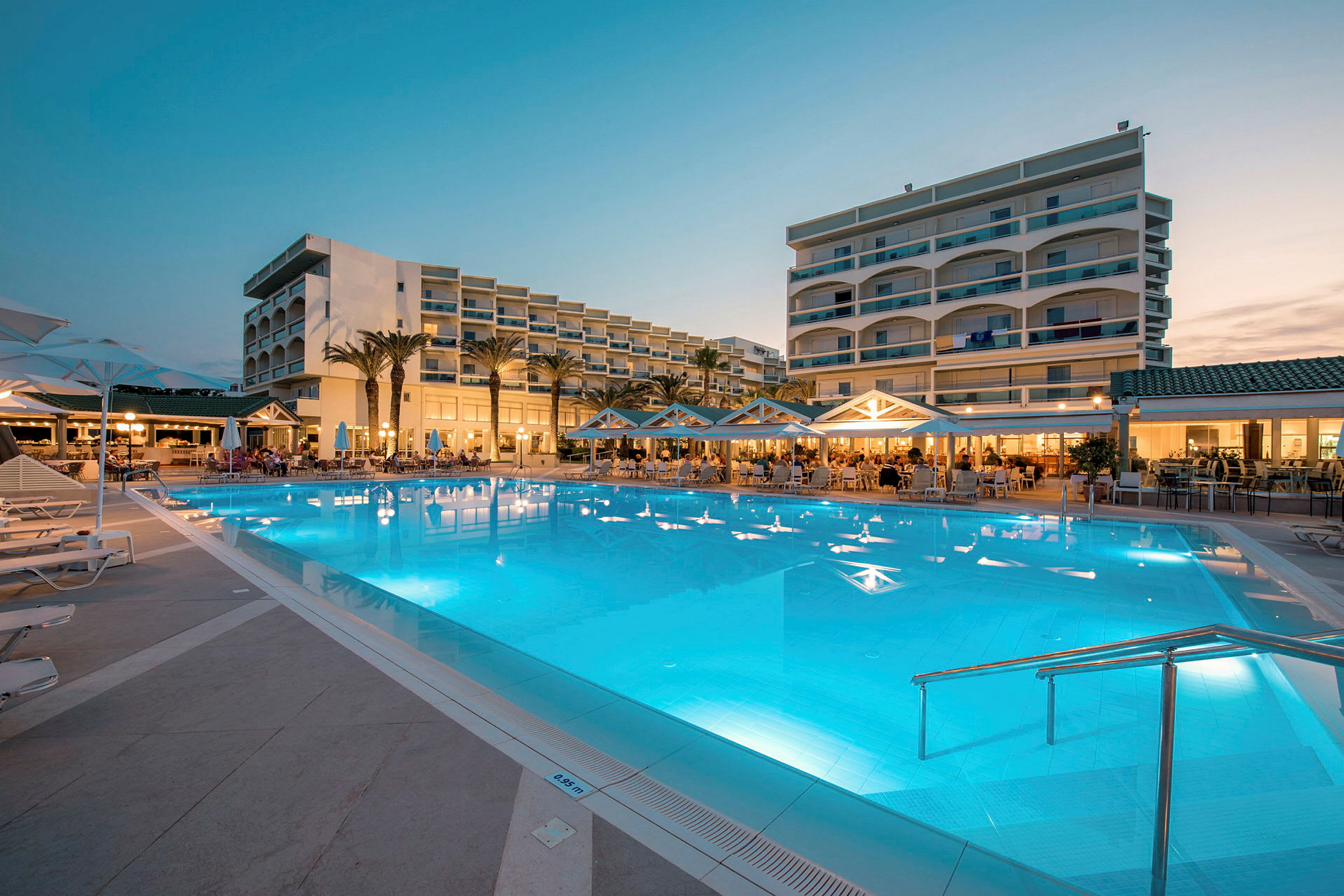 Udendørs pool med omkringliggende parasoller og hotelbygninger i baggrunden. (AI-genereret tekst)