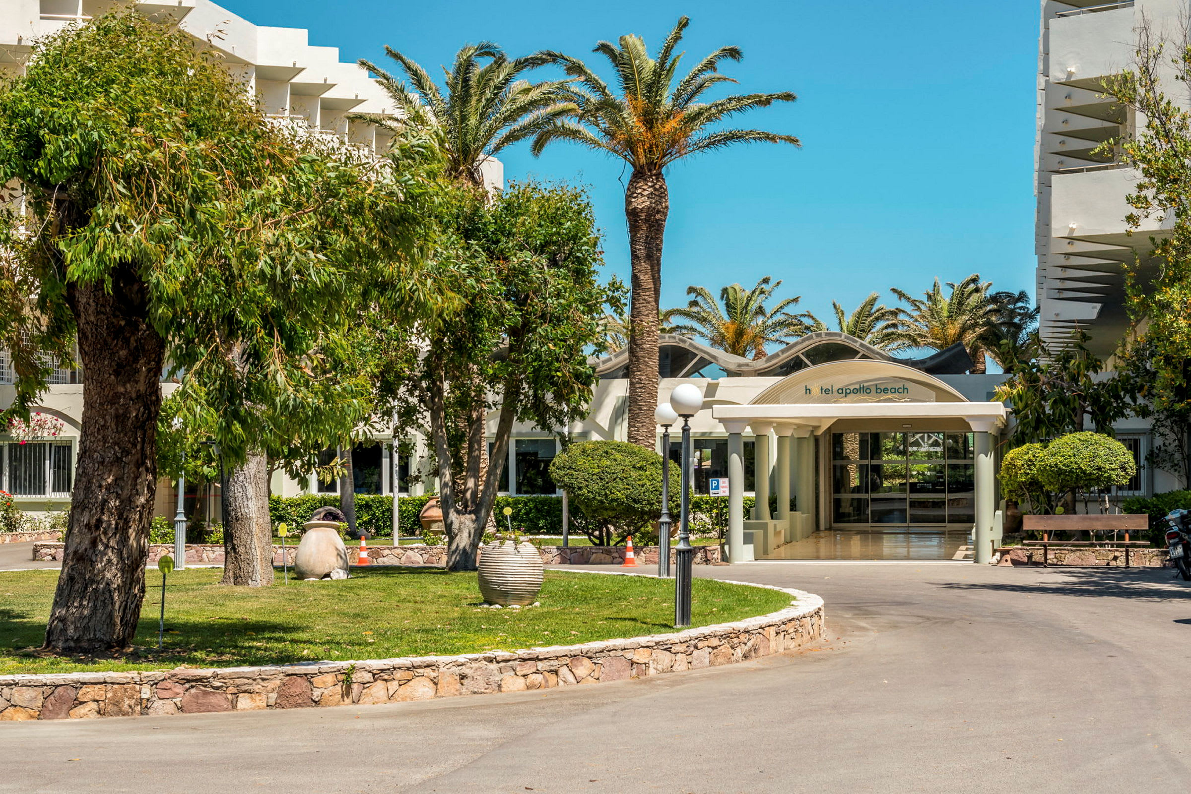 Indgangen til Hotel Apollo Beach omgivet af palmer og grønt. (AI-genereret tekst)