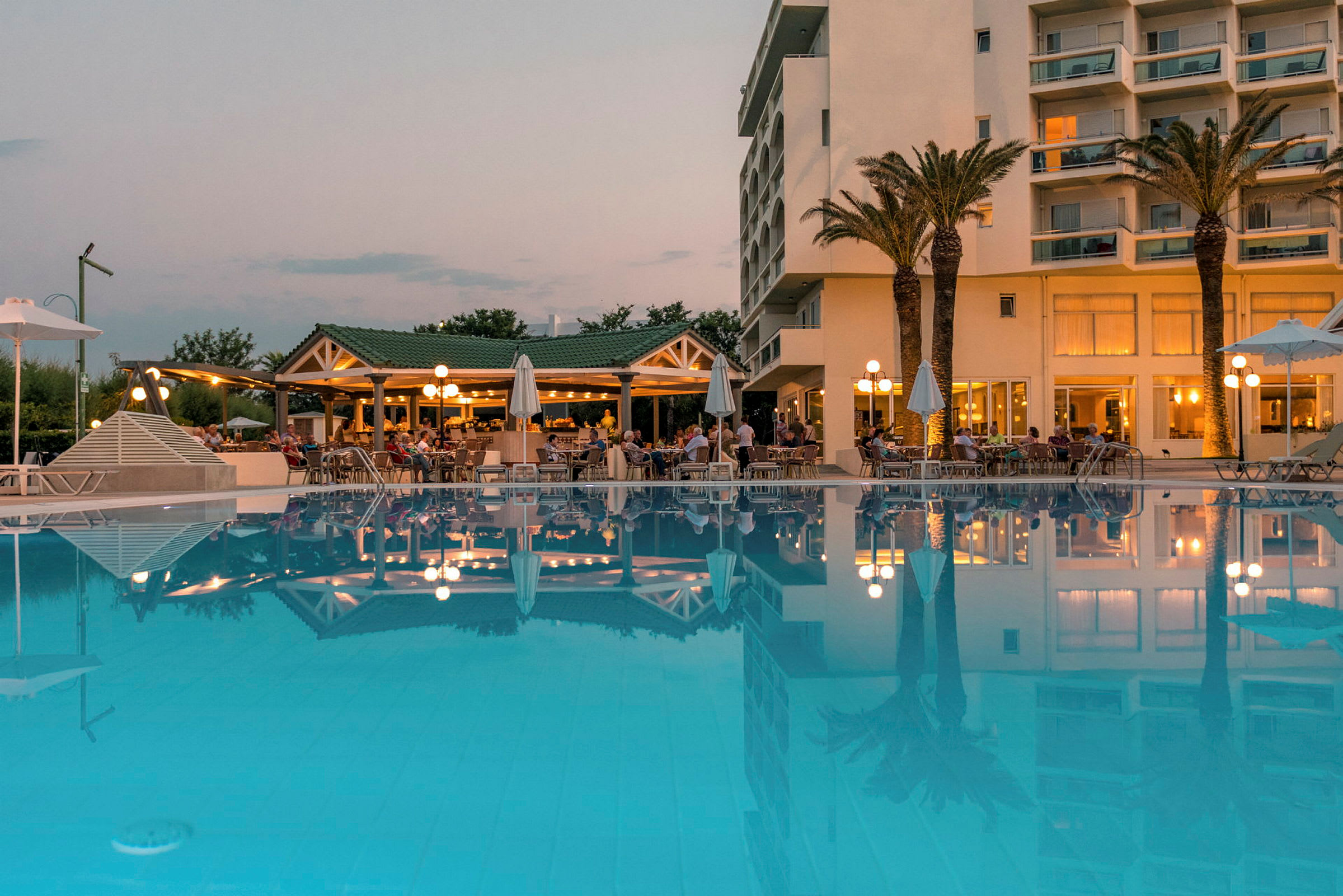 Hotelbygning med palmer og oplyst restaurant ved en stor pool. (AI-genereret tekst)