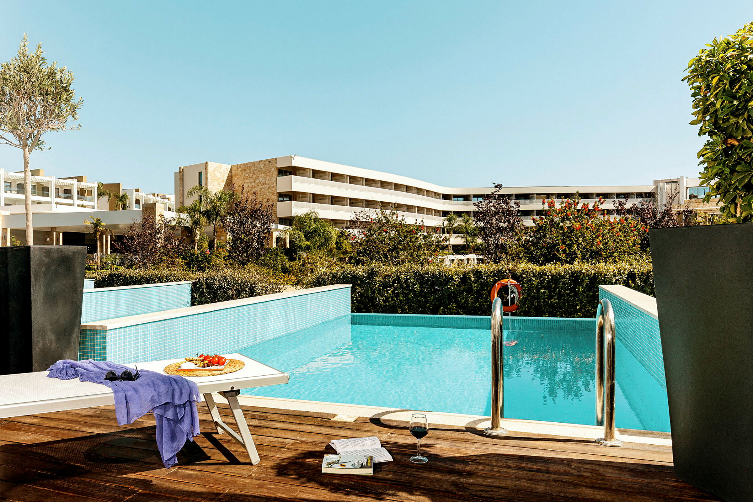 Udendørs område med pool, solseng, bord med frugt og glas samt hotelbygninger i baggrunden. (AI-genereret tekst)