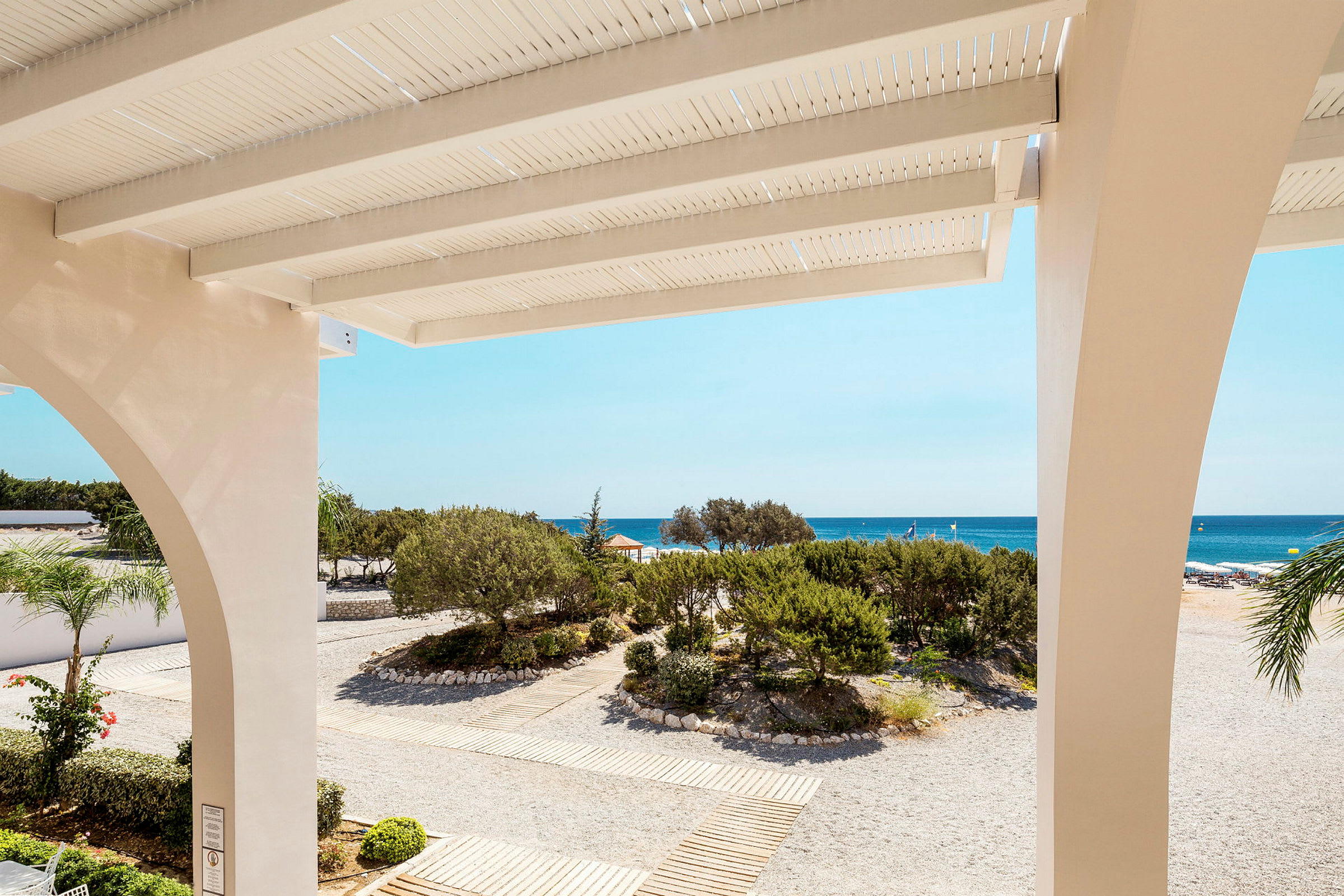 Udsigt fra en terrasse mod en strand med sand og grønne områder samt havet i baggrunden. (AI-genereret tekst)