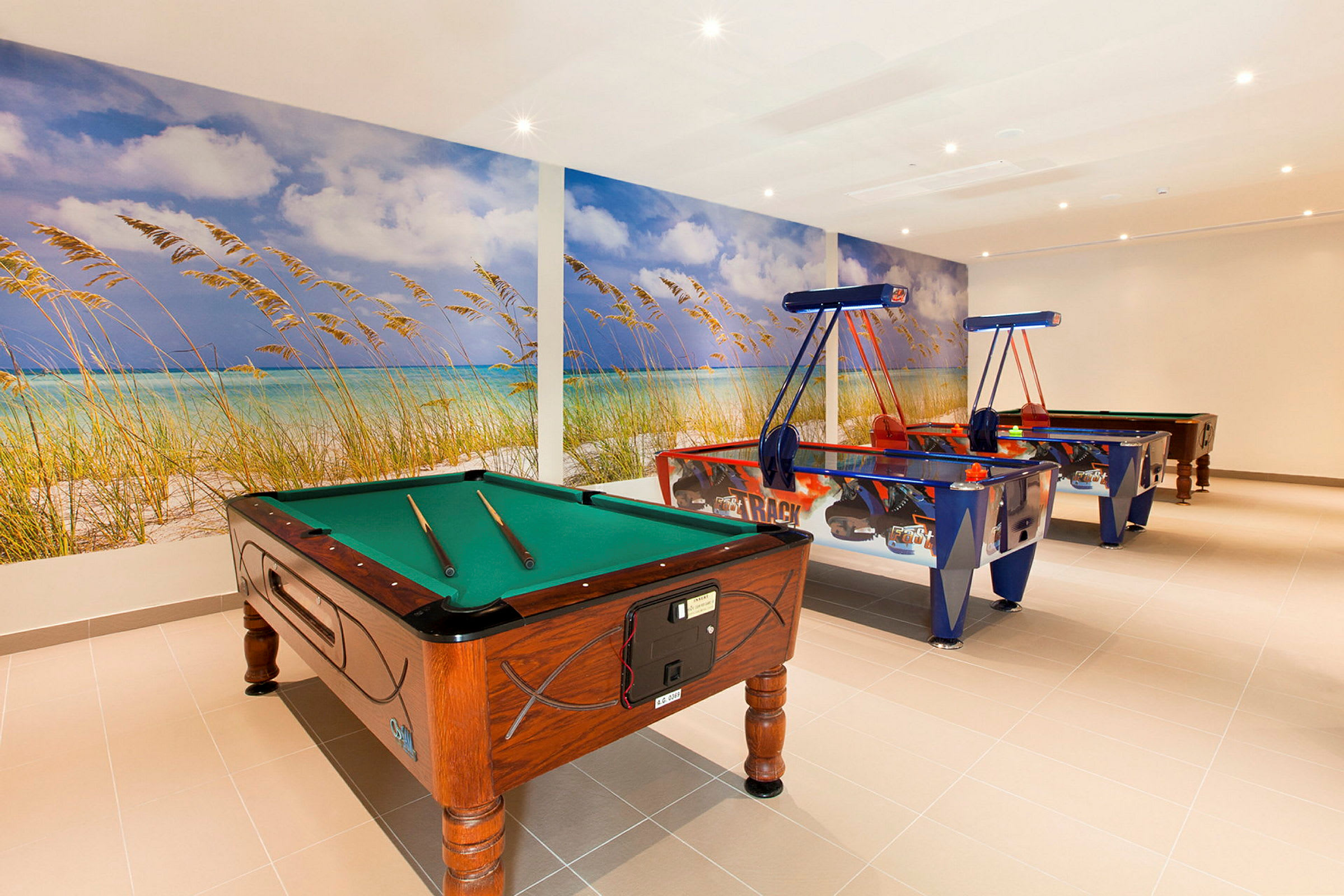 Et rum med et billiardbord og to airhockeyborde, med en vægmaleri af en strand som baggrund. (AI-genereret tekst)
