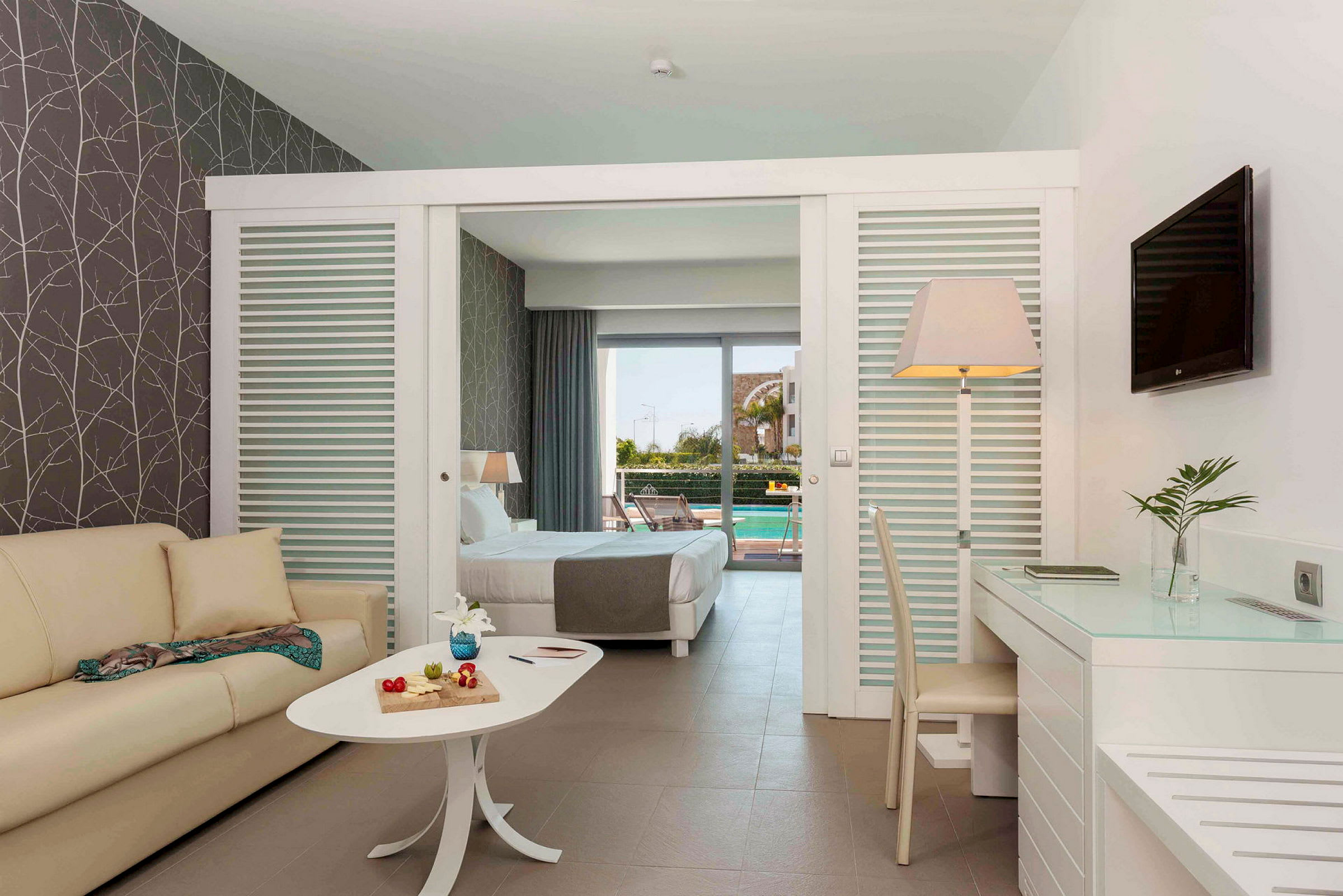Et hotelværelse med sofa, bord med dekorative genstande, skrivebord og stol. Sengen i baggrunden fører til en terrasse med udsigt over en pool. (AI-genereret tekst)