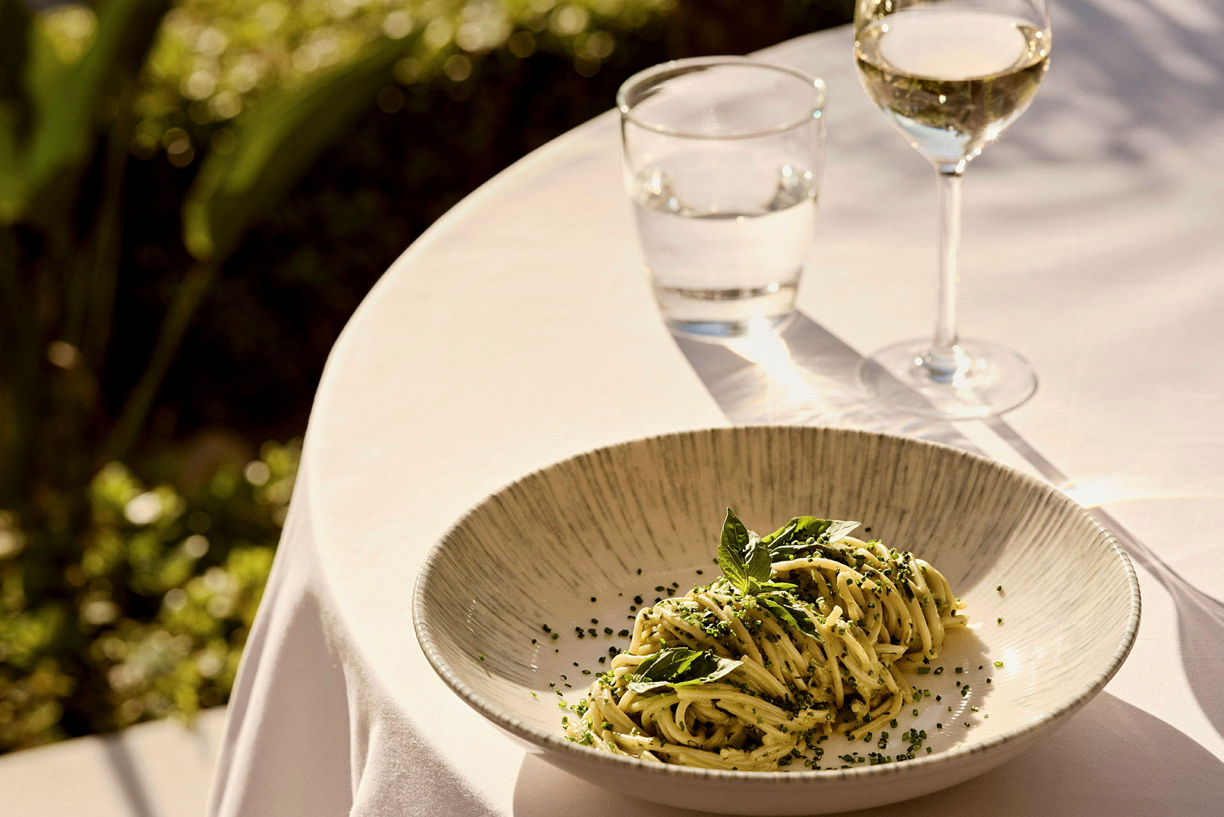 En tallerken med pasta dekoreret med urter, placeret på et bord dækket med hvid dug, ved siden af et glas vin og et glas vand. (AI-genereret tekst)