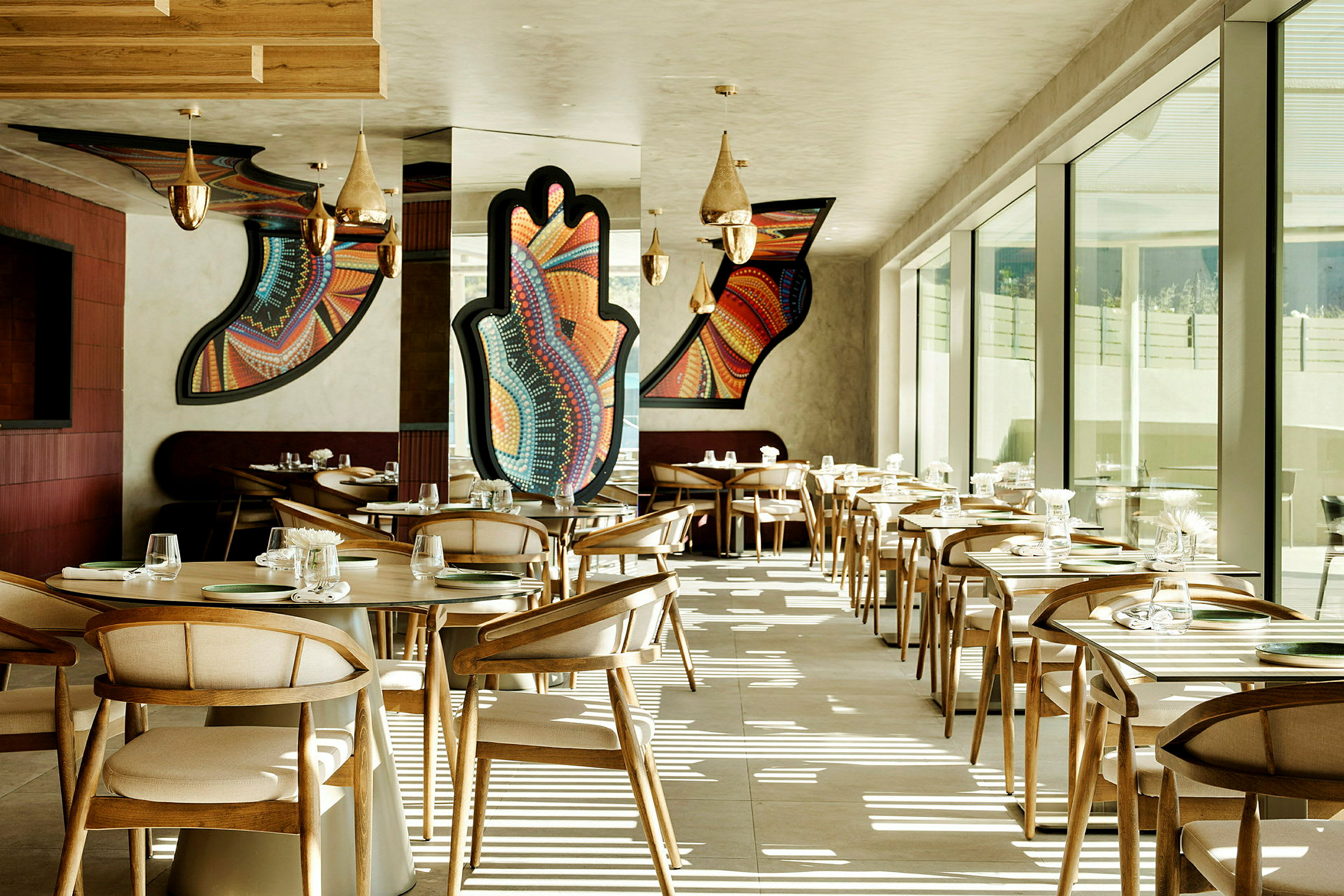 En moderne restaurant med træstole og borde. Væggene har farverige geometriske kunstværker. Store vinduer lader naturligt lys komme ind. (AI-genereret tekst)