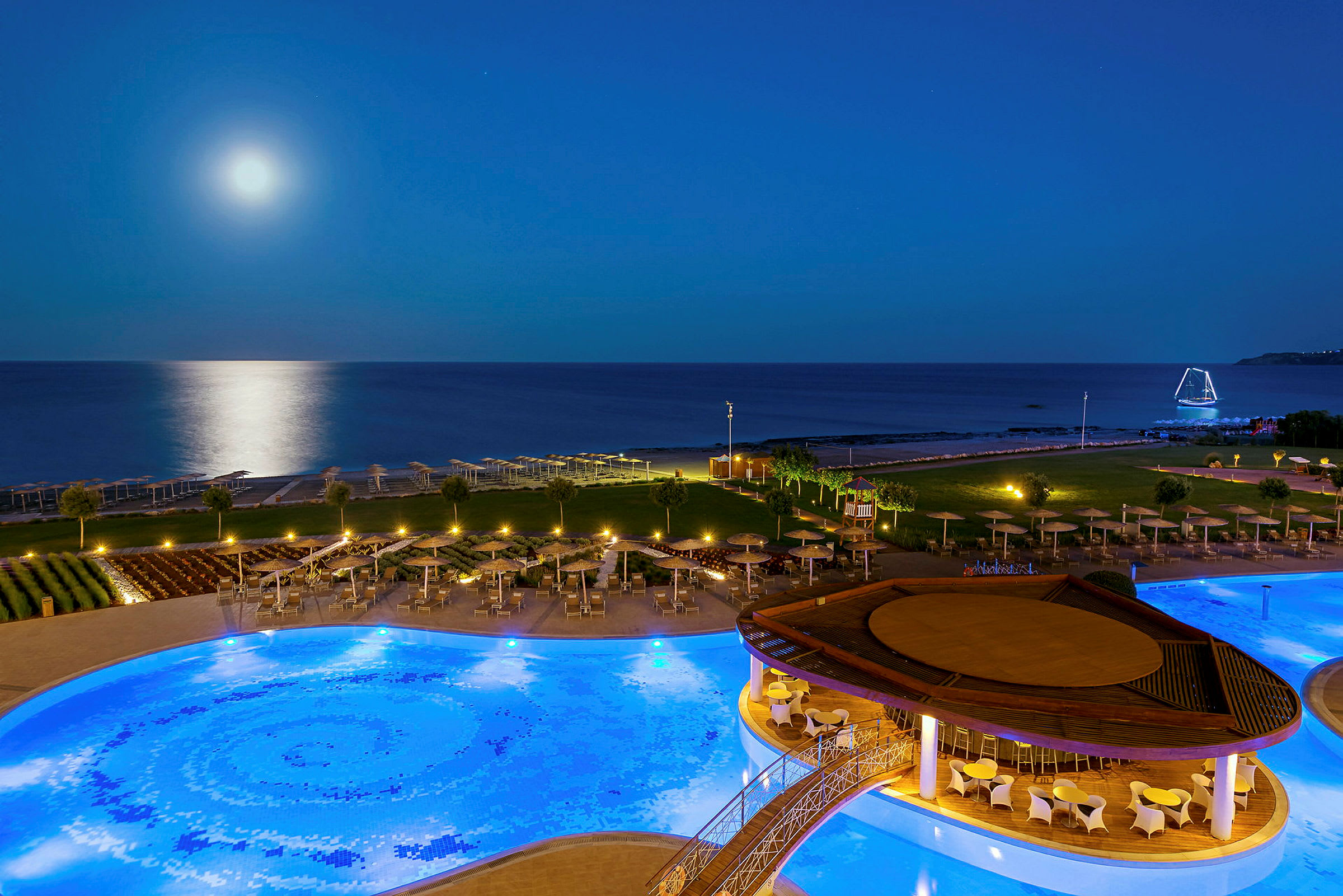 Aftenudsigt over en oplyst pool på en resort nær havet, med fuldmåne og refleksioner på vandet. (AI-genereret tekst)
