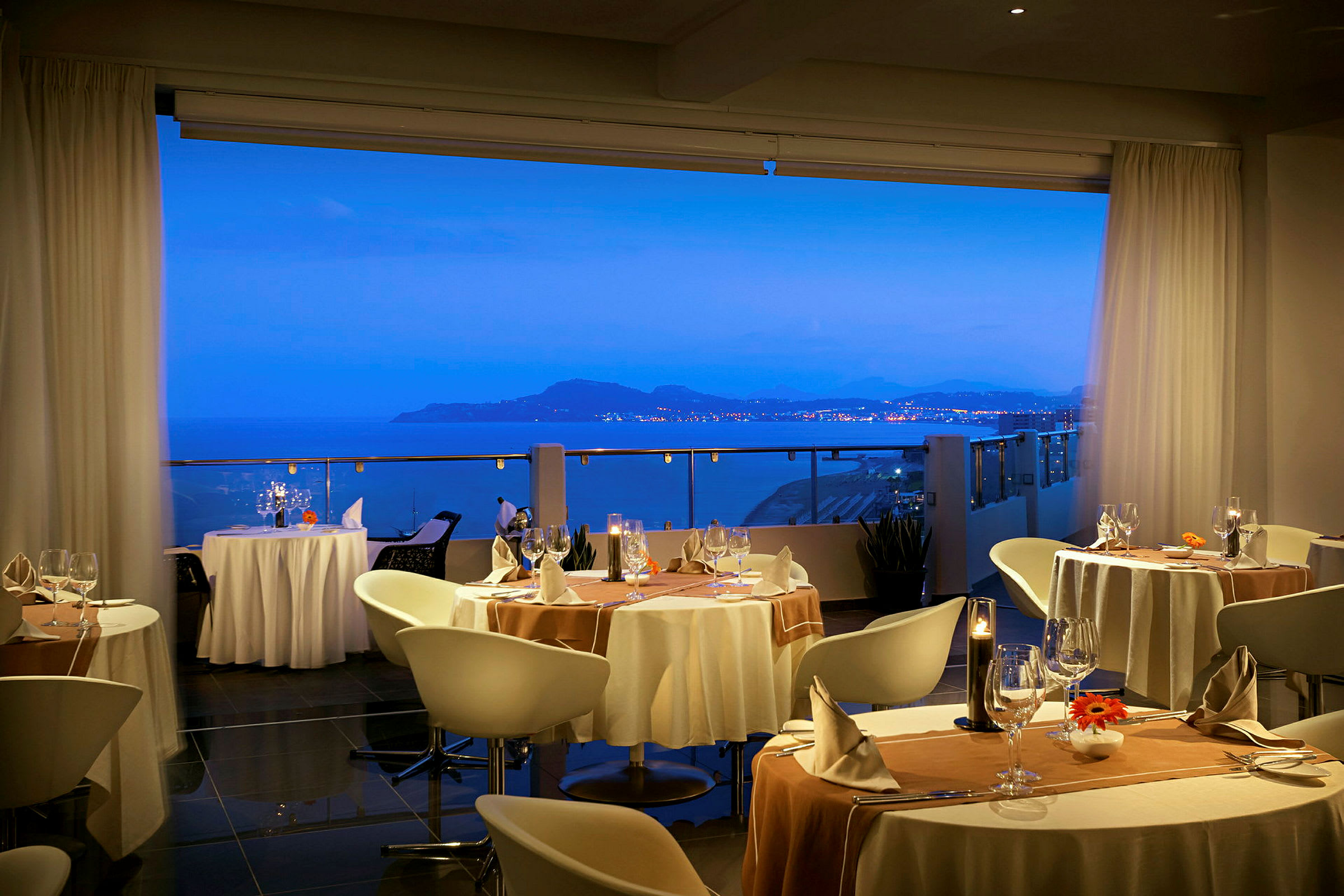 Udsigt over en havbugt med fjerne lys på land fra en elegant restaurantinteriør. (AI-genereret tekst)