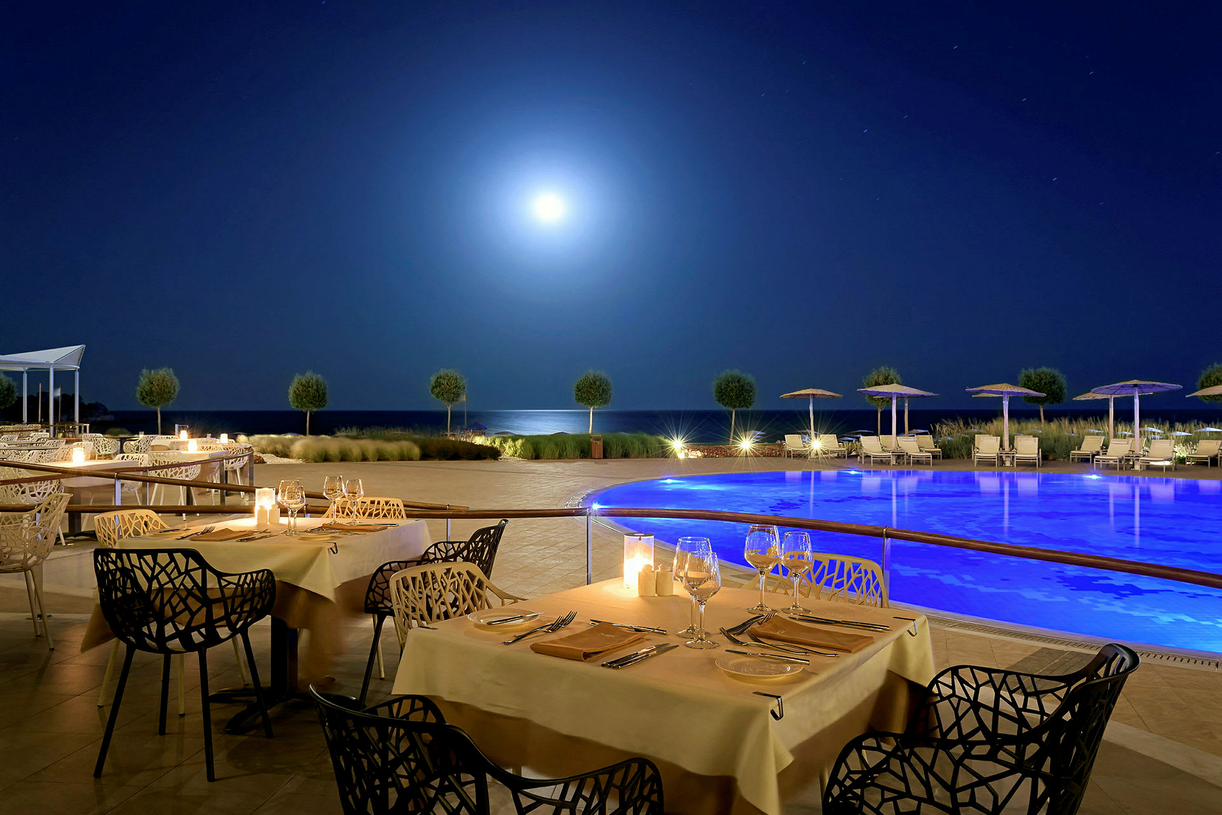 En udendørs restaurant med dækkede borde og en pool i nærheden under en stjerneklar nattehimmel. (AI-genereret tekst)