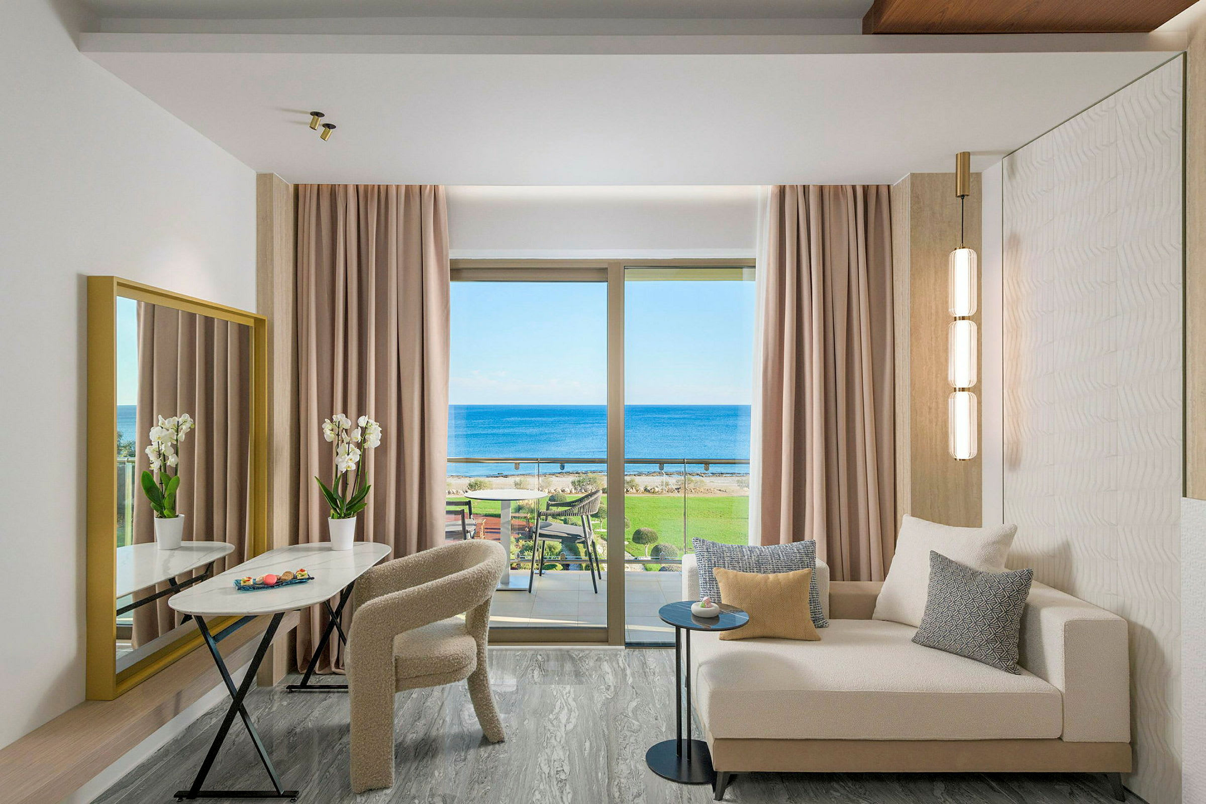 En moderne hotelsuite med sofa, bord, stole og udsigt over havet gennem et stort vindue. (AI-genereret tekst)