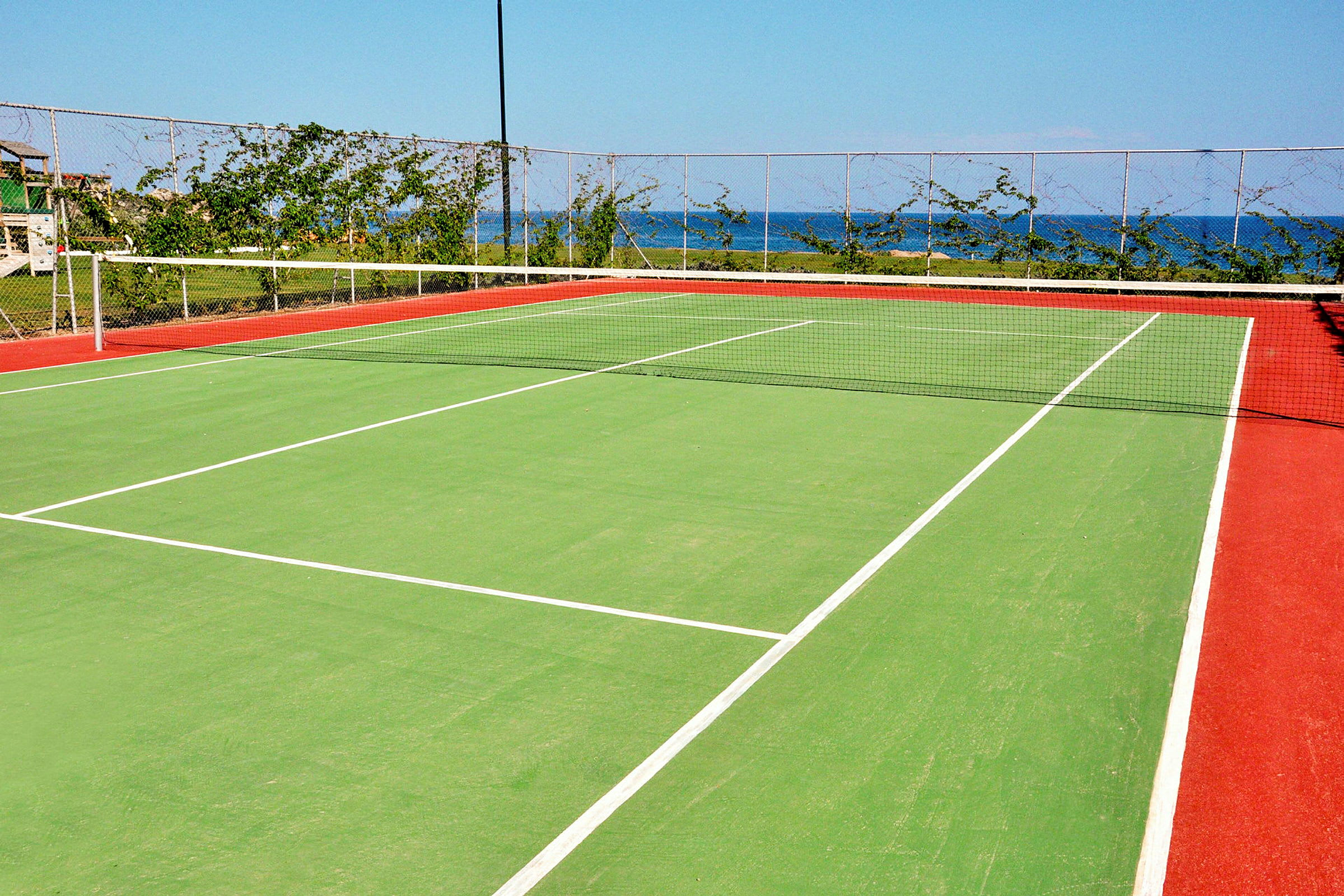 En tennisbane med grøn overflade og røde kanter, omgivet af et nethegn og grøn bevoksning, med havet synligt i baggrunden. (AI-genereret tekst)