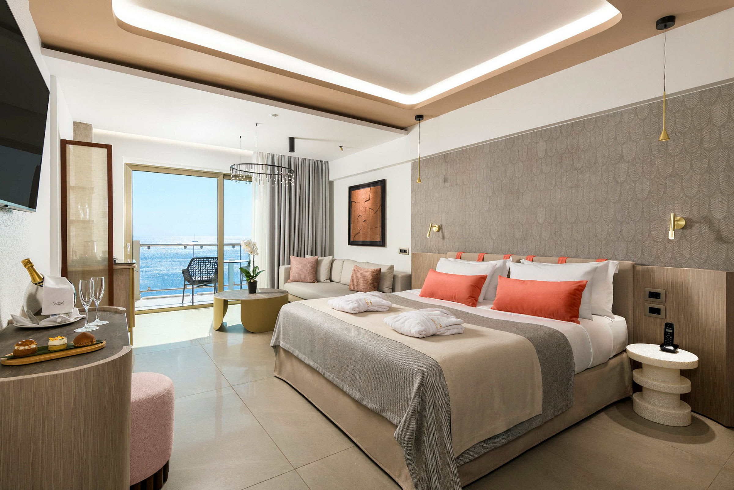 En moderne hotelsuite med redt seng med orange puder, siddegruppe og altan med havudsigt. (AI-genereret tekst)