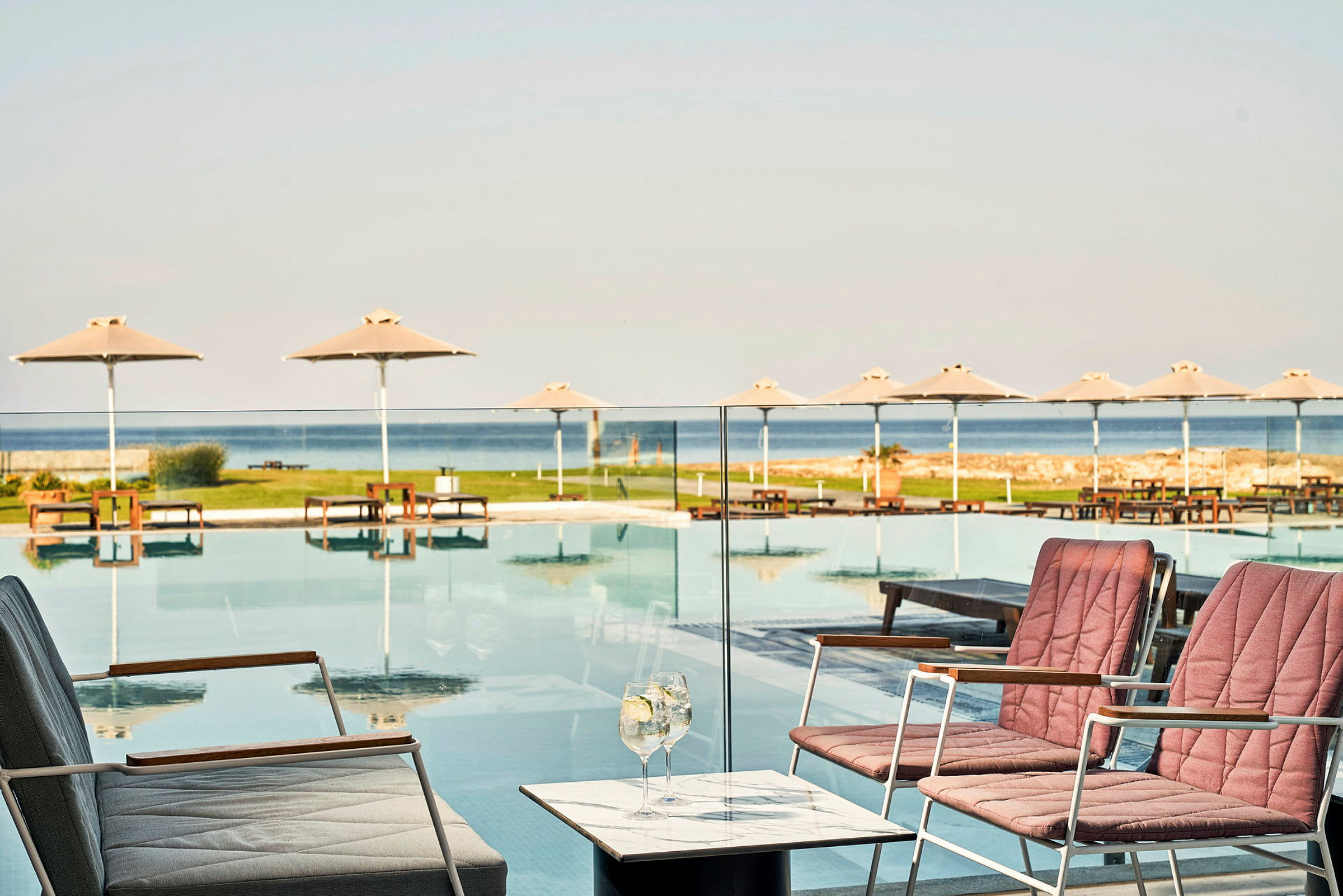 Udendørs poolområde med stole og borde i forgrunden, liggestole og parasoller ved poolen samt havet i baggrunden. (AI-genereret tekst)