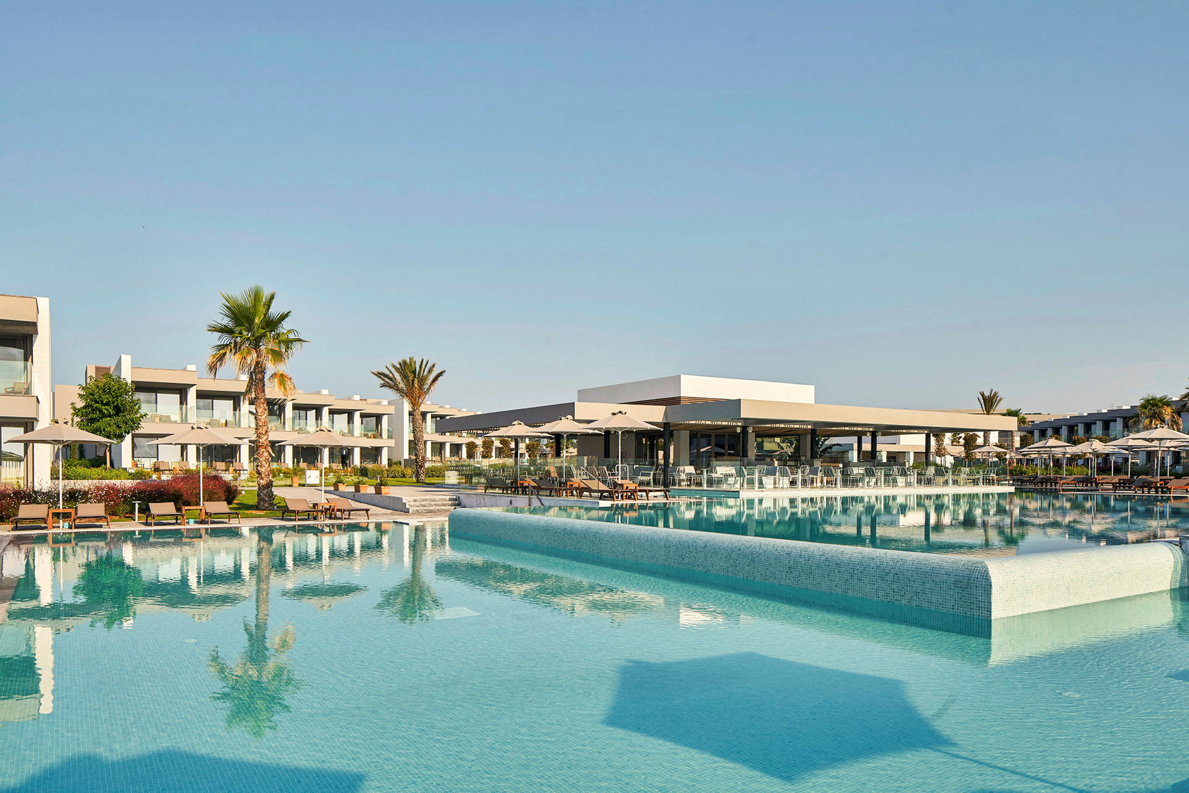Billede af en hotelpool med omkringliggende liggestole og bygninger. (AI-genereret tekst)