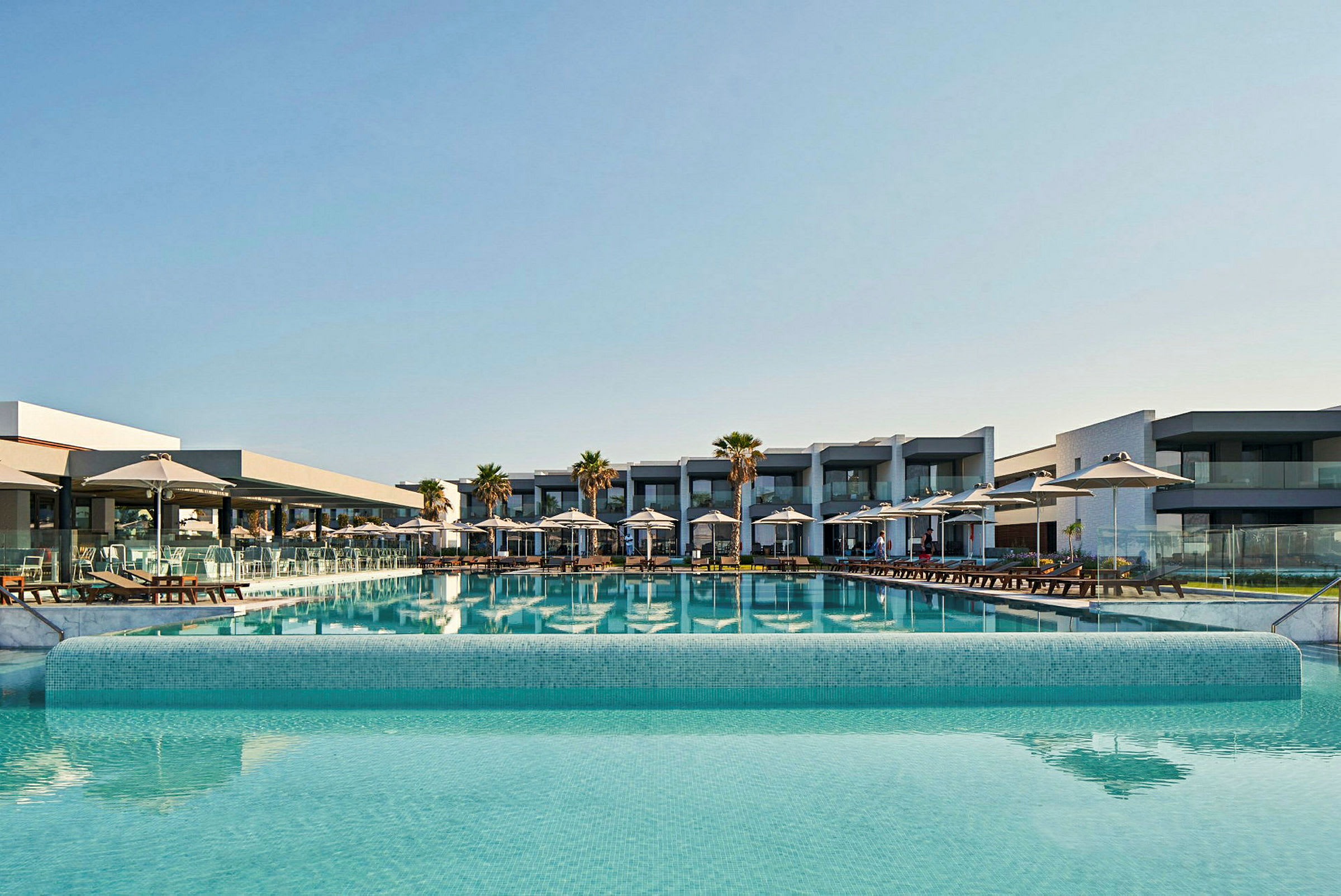 Moderne hotelbygning med stor pool og liggestole. (AI-genereret tekst)
