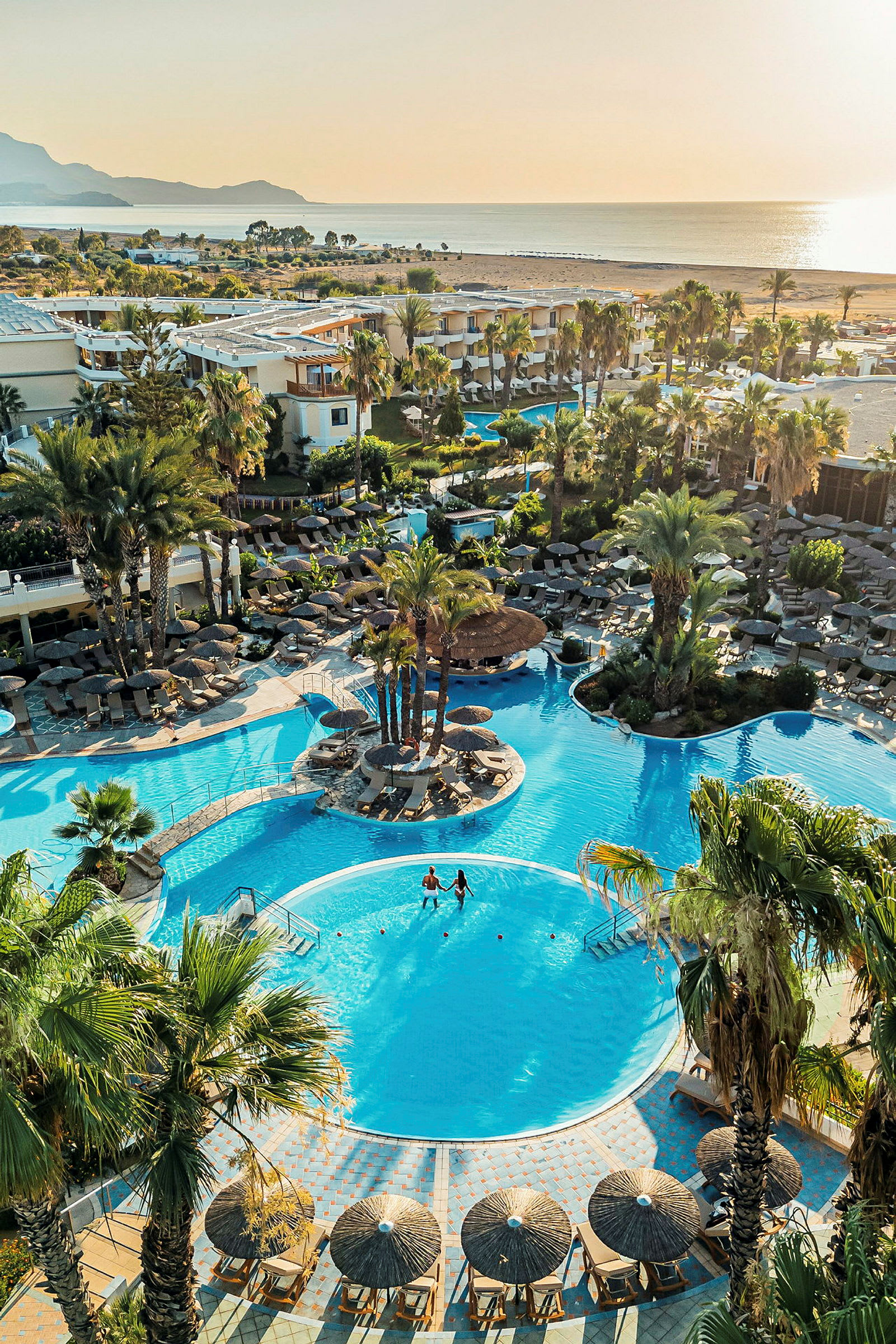 Hotelområde med pool omgivet af palmer og liggestole tæt på havet. (AI-genereret tekst)