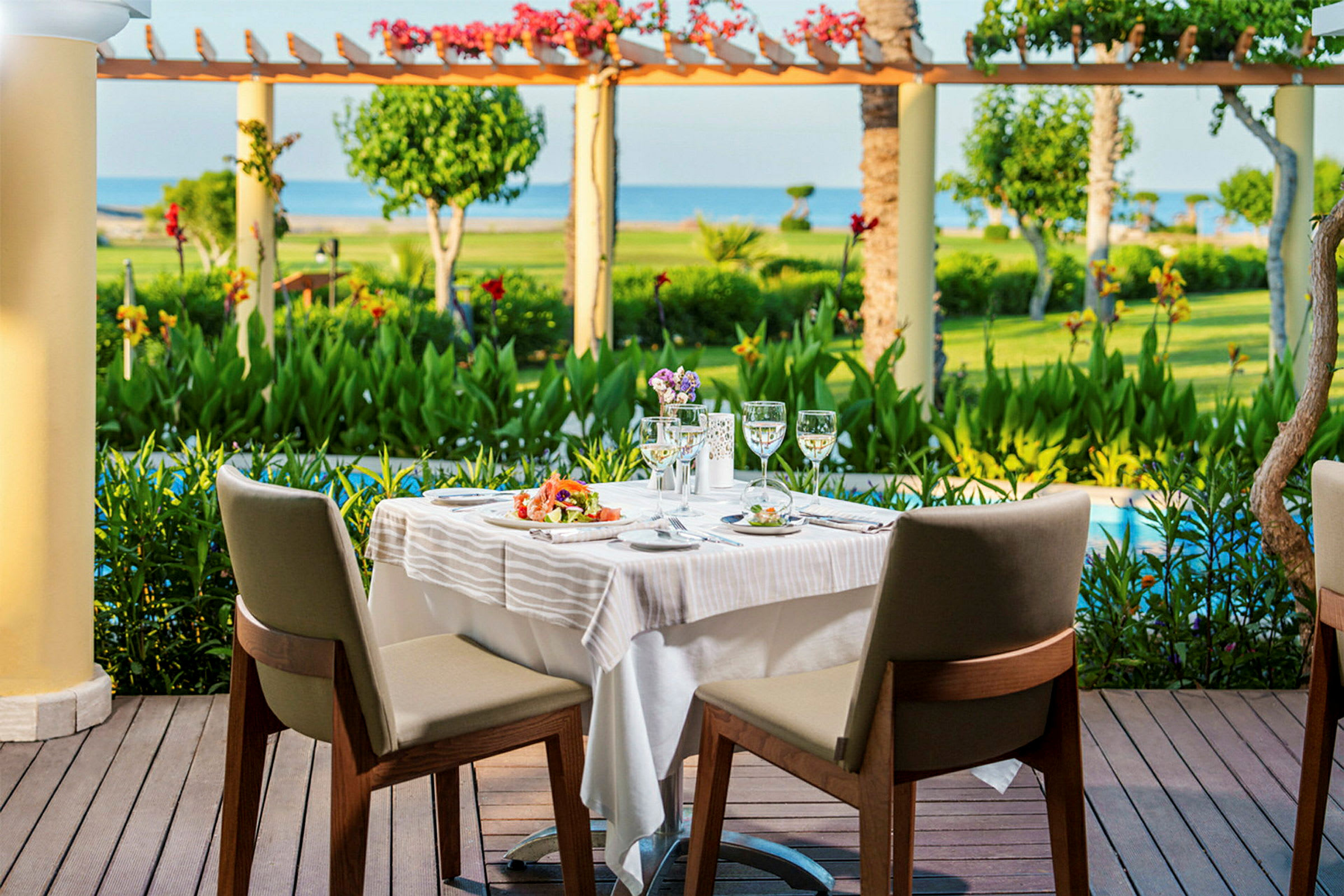 To spisebordsstole ved et dækket bord på en terrasse med udsigt over en have og havet. (AI-genereret tekst)